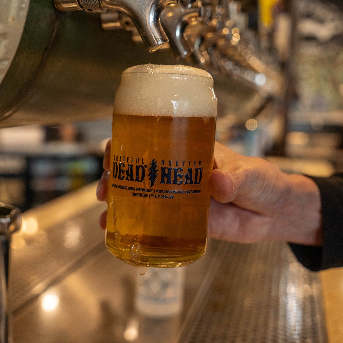 Freshly poured beer in Dogfish Head x Grateful Dead 2026 Record Store Day can glass with foamy head at brewery bar.