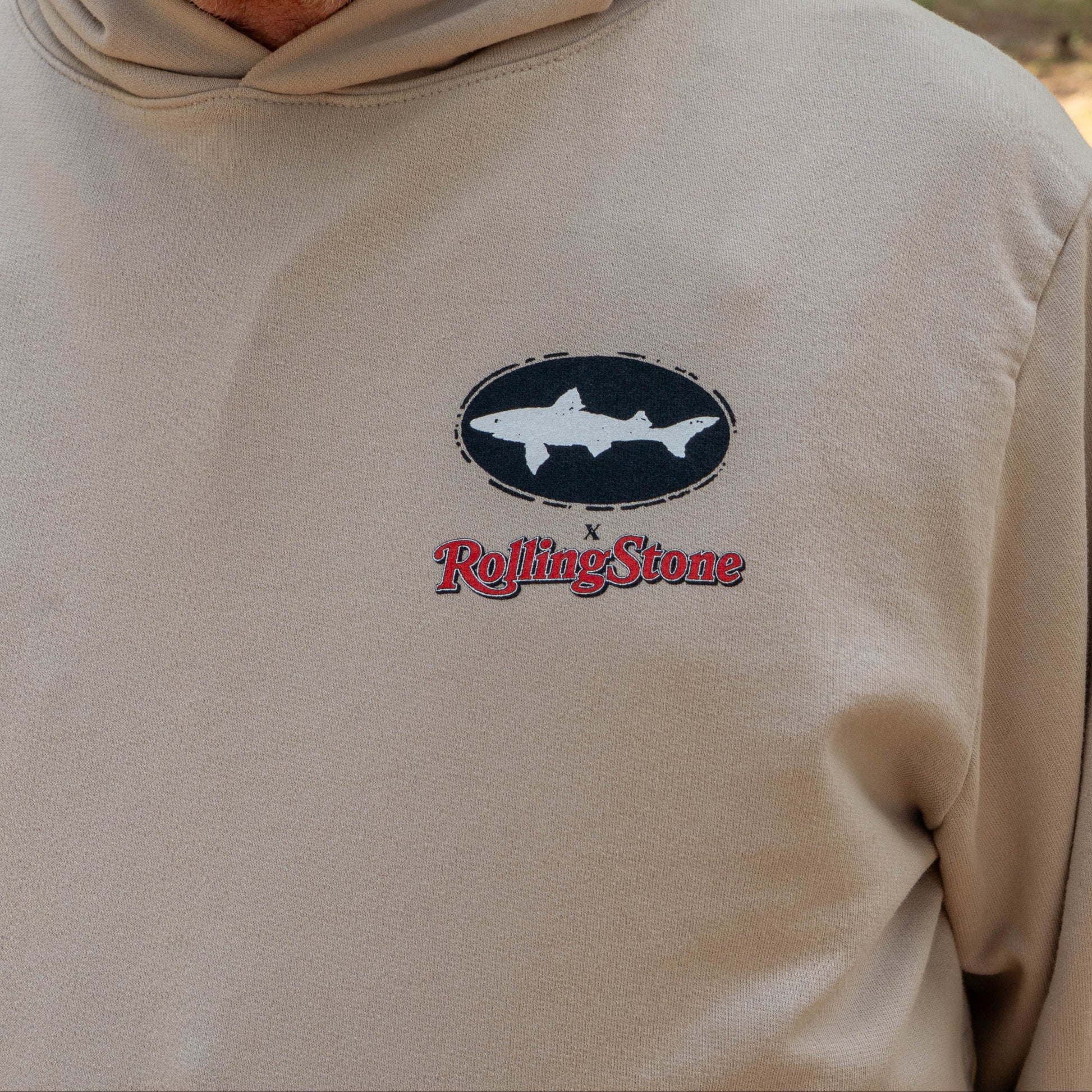 Person wearing Dogfish Head x Rolling Stone Mocha Hoodie  featuring a black oval Dogfish logo logo a white shark and red Rolling Stone logo