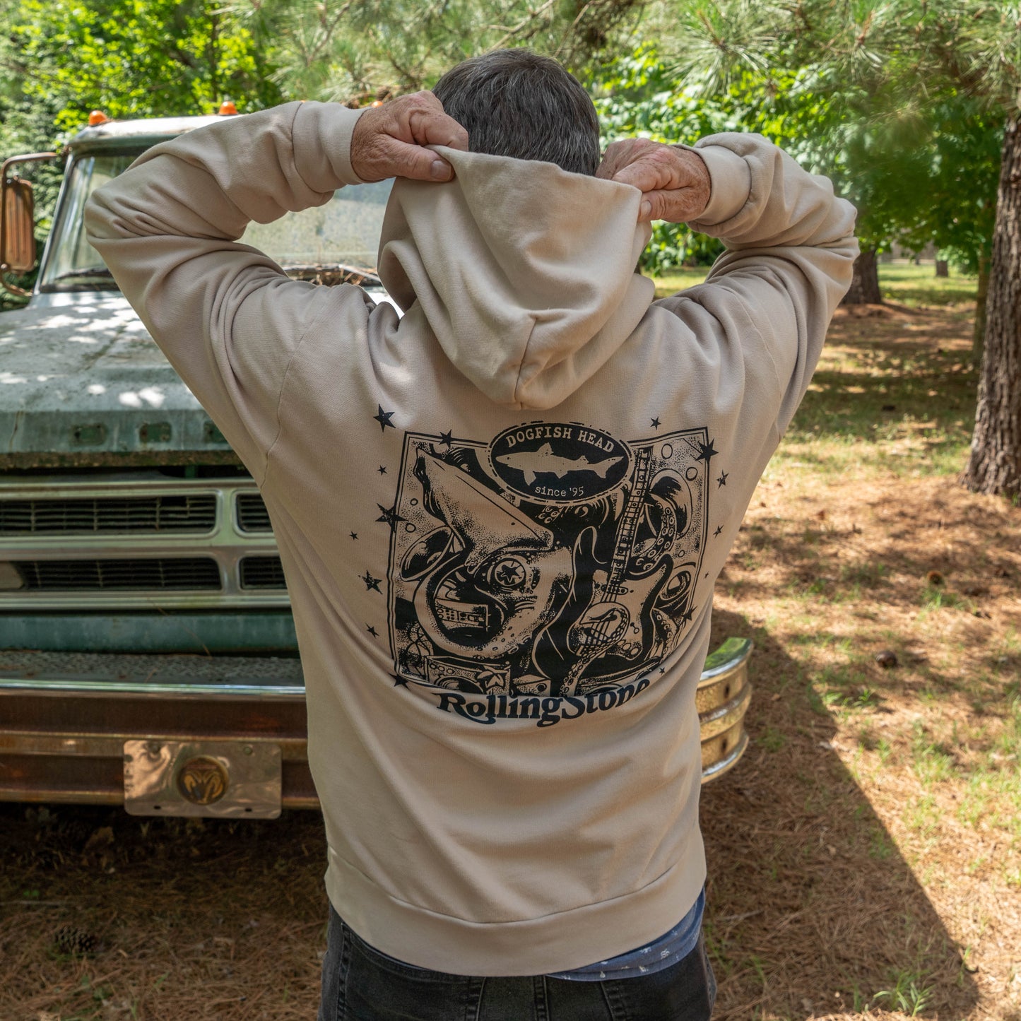 A person outdoors wearing a tan Dogfish Head x Rolling Stone Mocha Hoodie featuring a bold black squid and guitar graphic and logo on the back. 