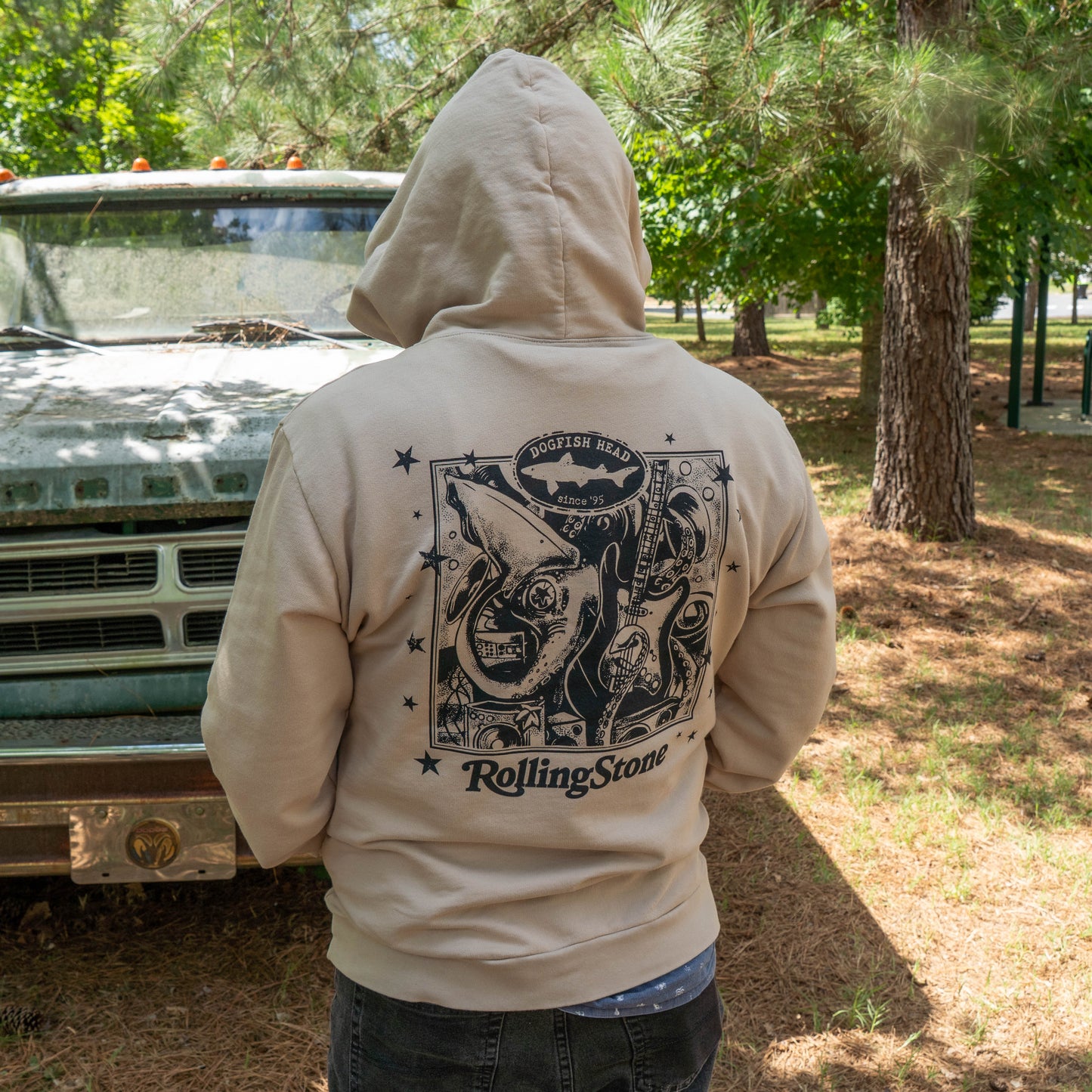 A person stands outdoors with their back to the camera, wearing the GO WEST T-SHIRT COMPANY Dogfish Head x Rolling Stone Mocha Hoodie near an old truck, surrounded by trees and sunlight.