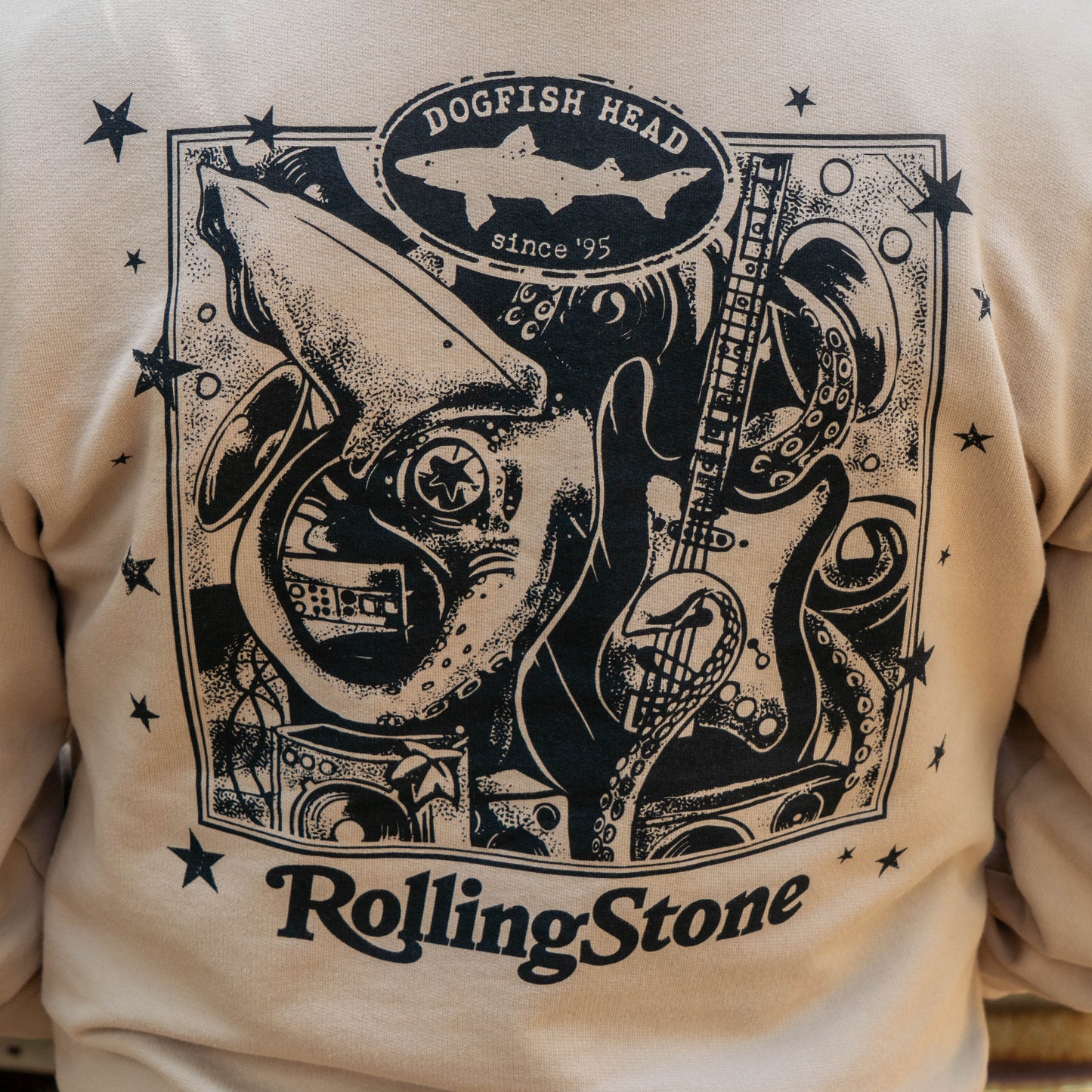 The Dogfish Head x Rolling Stone Mocha Hoodie by GO WEST T-SHIRT COMPANY features a black graphic with a shark, guitar, and abstract art plus “Dogfish Head since ’95” at the top and “Rolling Stone” at the bottom.