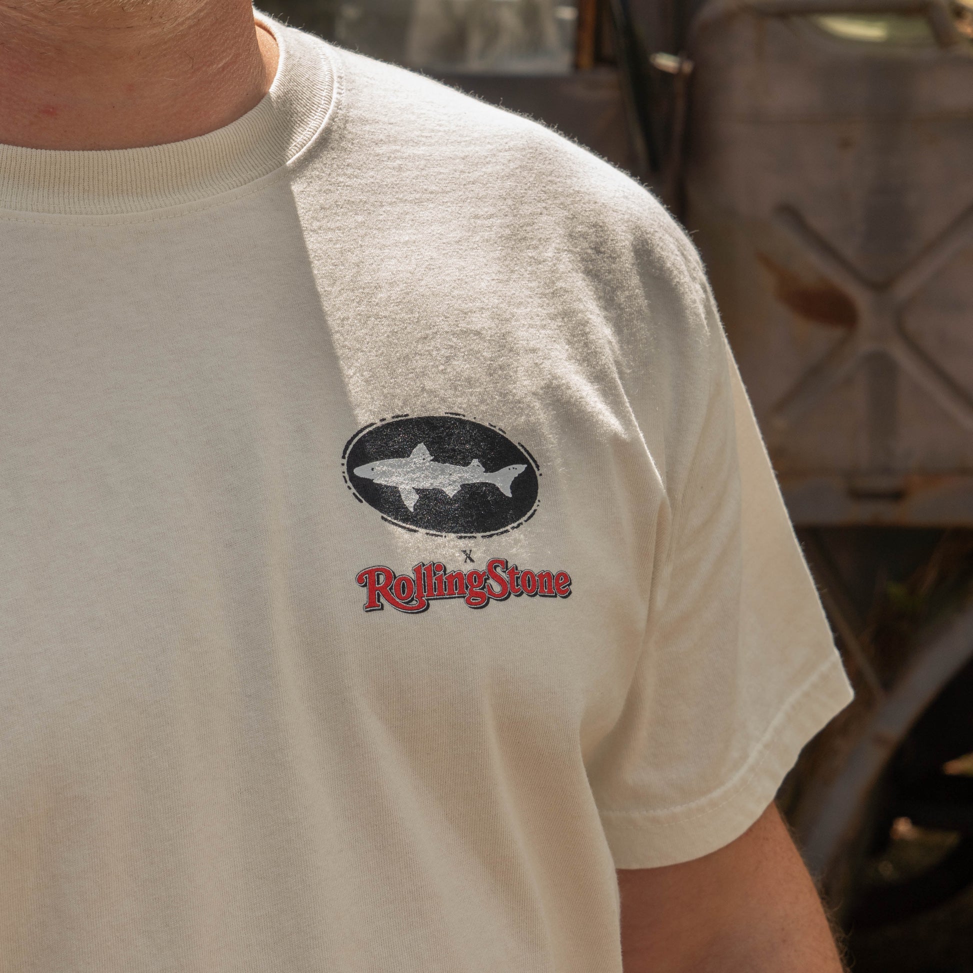 A person wearing the Dogfish Head x Rolling Stone Oatmeal Tee Shirt featuring a black Dogfish Head oval shark logo above red Rolling Stone logo