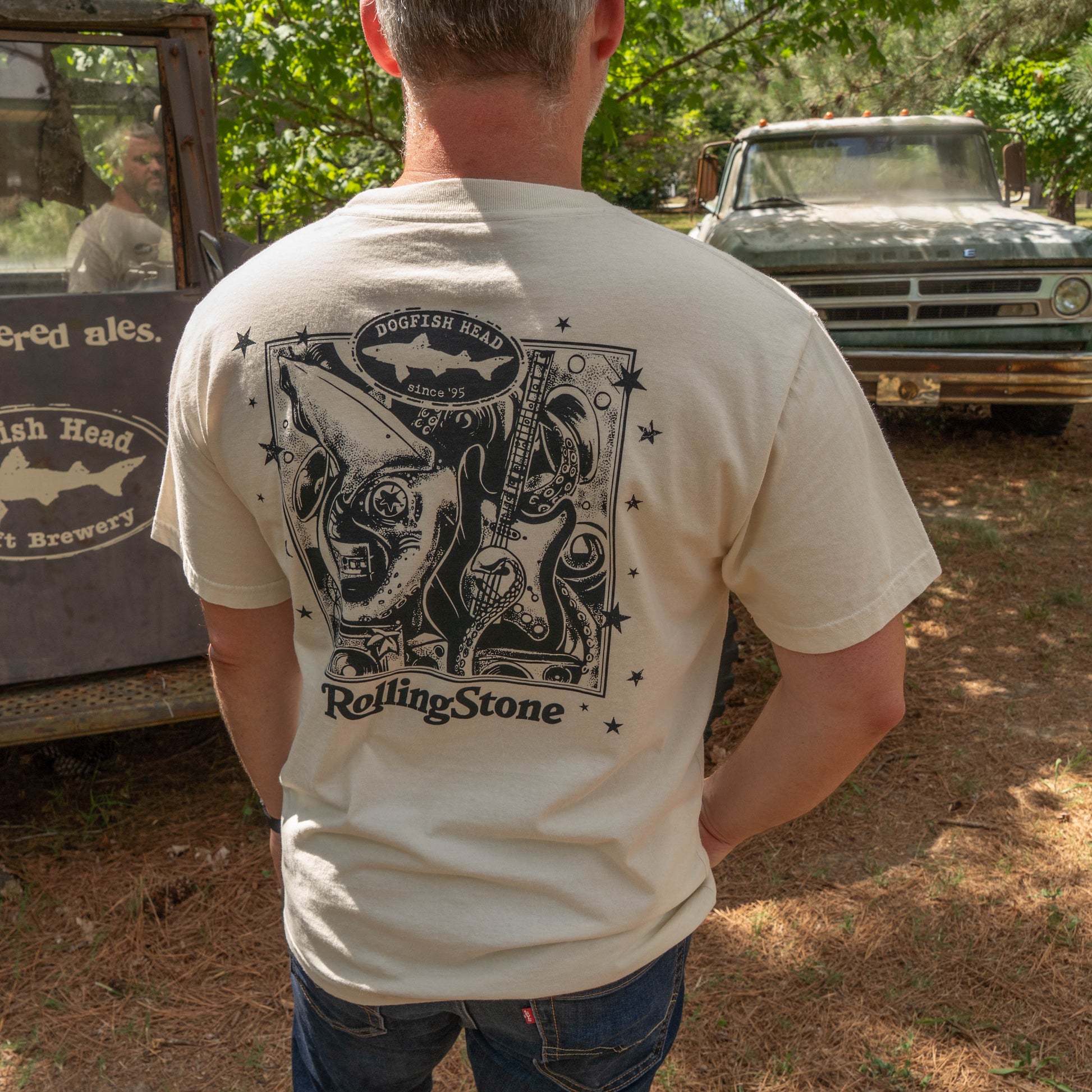 A man outdoors, back turned, wears a Dogfish Head x Rolling Stone Oatmeal Tee Shirt with a detailed Squid, electric guitar, and star graphics 
