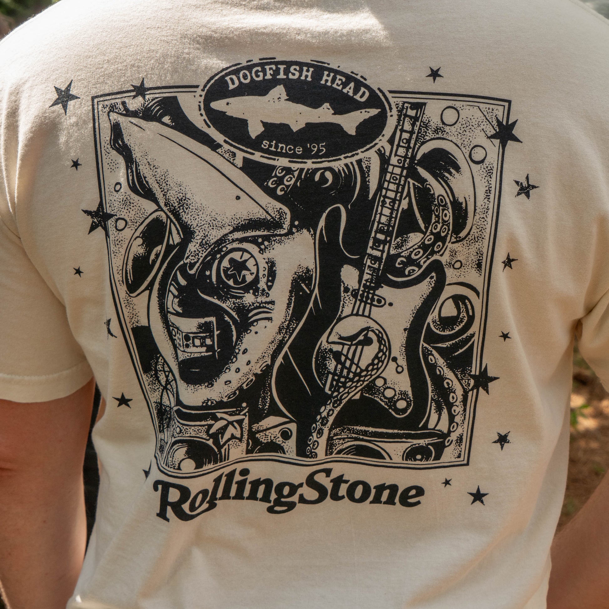 The back of the cream-colored Dogfish Head x Rolling Stone Oatmeal Tee Shirt  features a black graphic with a shark, guitar, stars, and "Dogfish Head since 95" and "Rolling Stone.