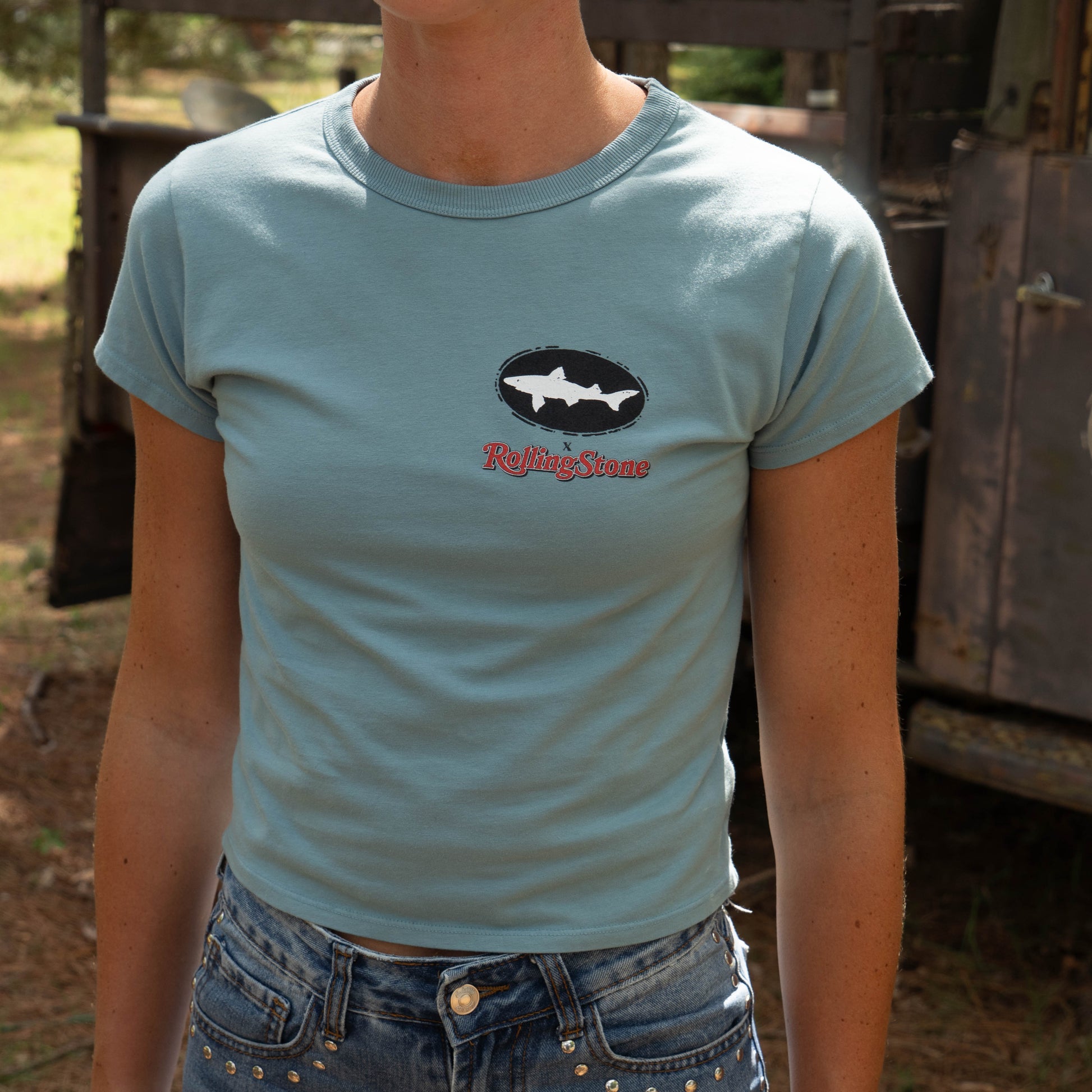 Outdoors, someone wears the Dogfish Head x Rolling Stone Women's Slate Blue Baby Tee Shirt with blue jeans 