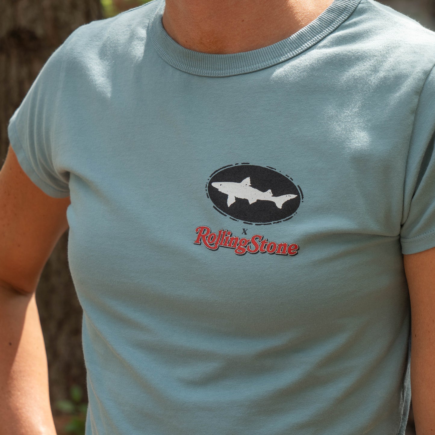 A person wearing a Dogfish Head x Rolling Stone Women's Slate Blue Baby Tee Shirt  with a black Dogfish Head logo and a red Rolling Stone logo on the left chest