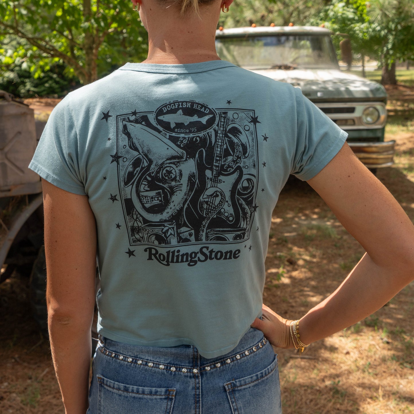 A woman in a Dogfish Head x Rolling Stone Women's Slate Blue Baby Tee Shirt with black detailed back graphics of a squid, electric guitar, stars, and bubbles. 