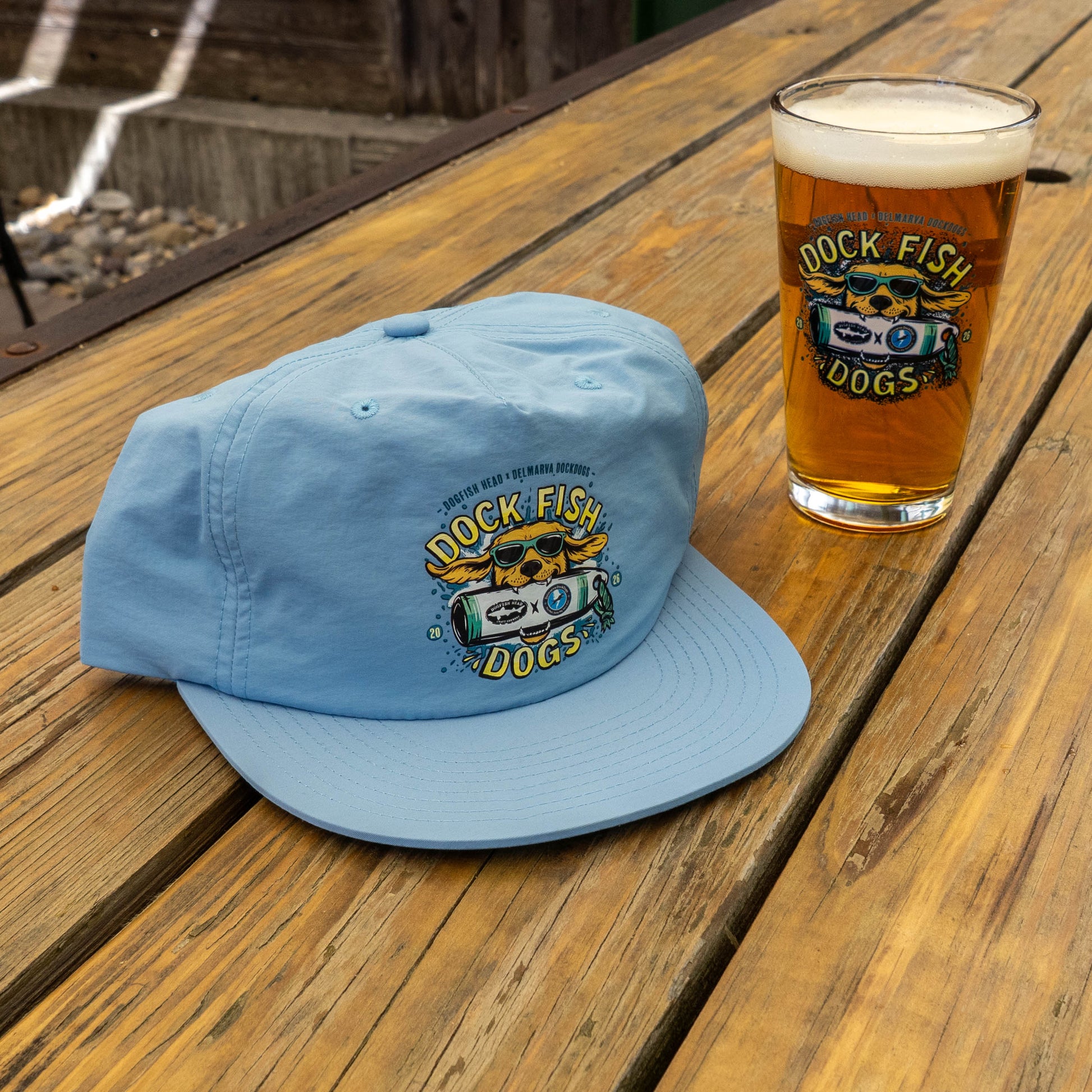 The Dock Dogs Carolina Blue Surf Hat by GO WEST T-SHIRT COMPANY rests on a wooden table beside a pint glass of beer, both sporting the Dock Fish Dogs logo.