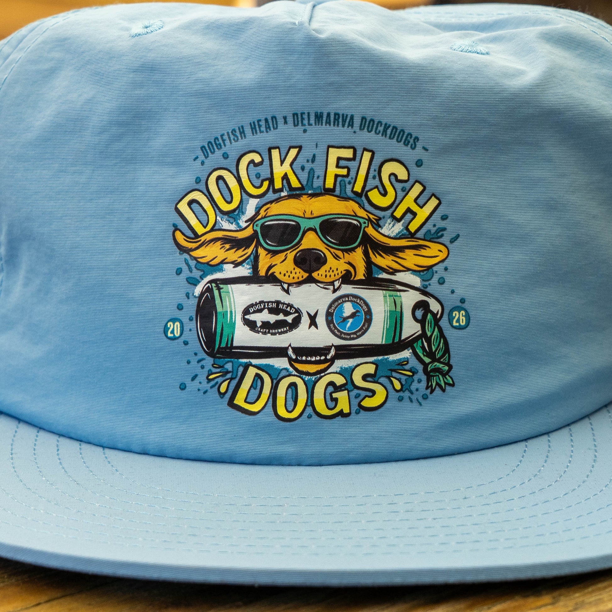 The Dock Dogs Carolina Blue Surf Hat by GO WEST T-SHIRT COMPANY features a dog with sunglasses holding a surfboard, plus “Dock Fish Dogs” and “Dogfish Head x Delmarva Dockdogs” text above and around the logo.