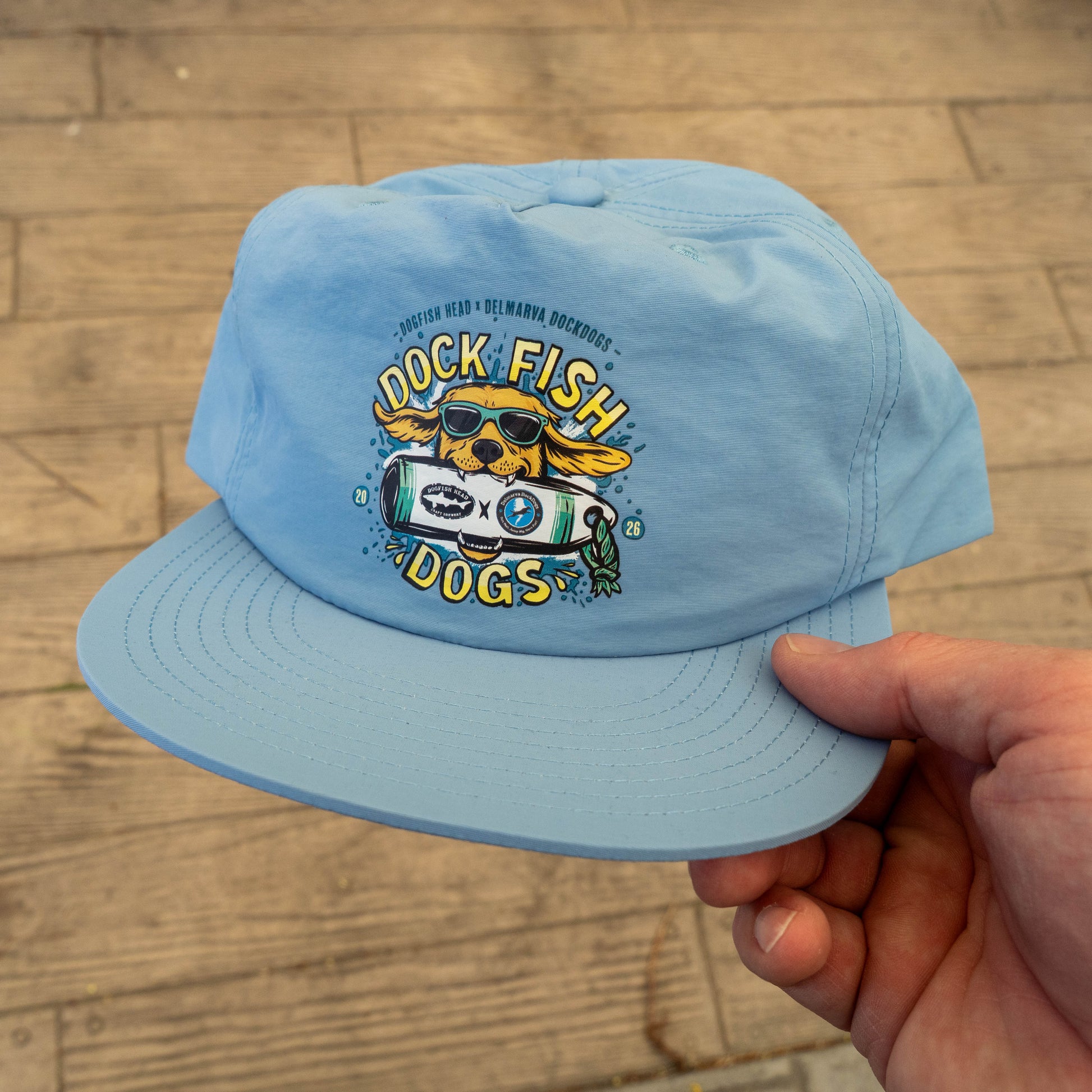 A hand holds the Dock Dogs Carolina Blue Surf Hat by GO WEST T-SHIRT COMPANY, featuring a cartoon dog in sunglasses and a fish. Made from recycled nylon, this surf cap adds playful style for fans of fun beachwear.