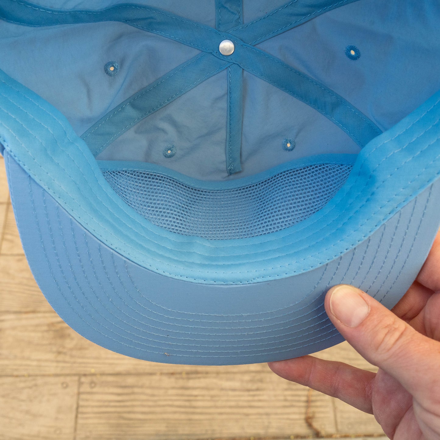 A close-up of a Dock Dogs Carolina Blue Surf Hat by GO WEST T-SHIRT COMPANY shows the interior stitching, mesh lining, and a hand holding the brim above a wooden surface.