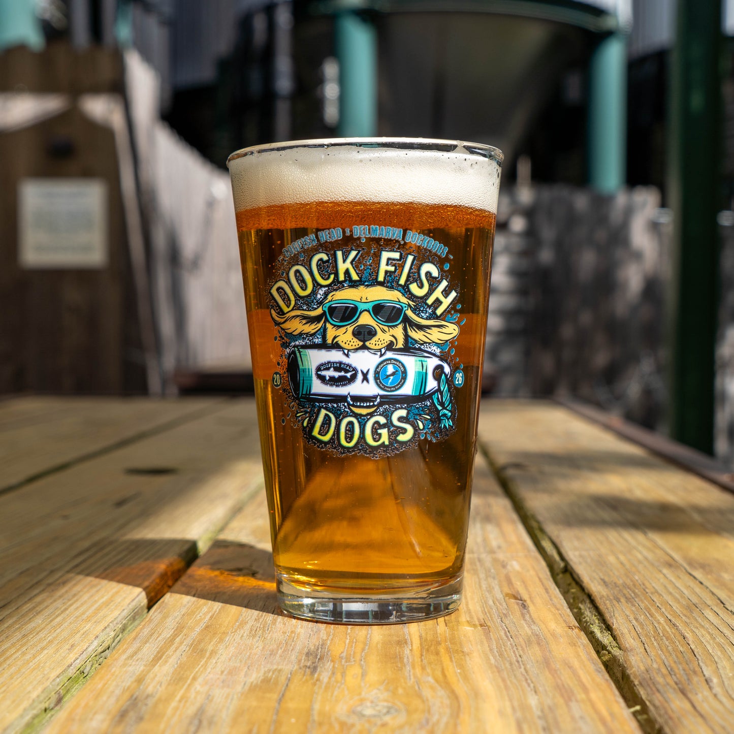 Pint glass with a graphic of a golden retriever wearing sunglasses biting into Dogfish Head x Delmarva Dock Dogs plushie. "Dog Fish Dogs" written in yellow 