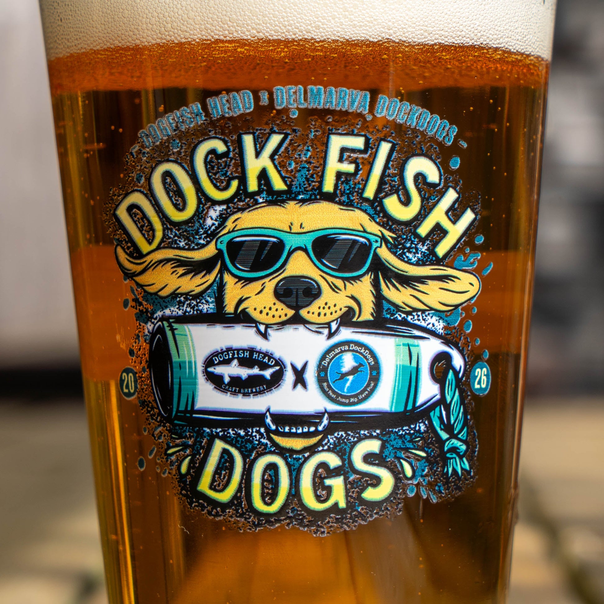 Pint glass with a graphic of a golden retriever wearing sunglasses biting into Dogfish Head x Delmarva Dock Dogs plushie. "Dog Fish Dogs" written in yellow 