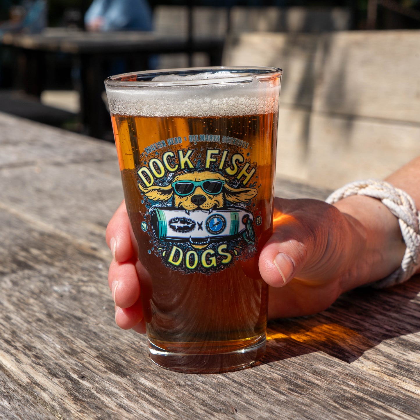 Man holding pint glass with a graphic of a golden retriever wearing sunglasses biting into Dogfish Head x Delmarva Dock Dogs plushie. "Dog Fish Dogs" written in yellow 
