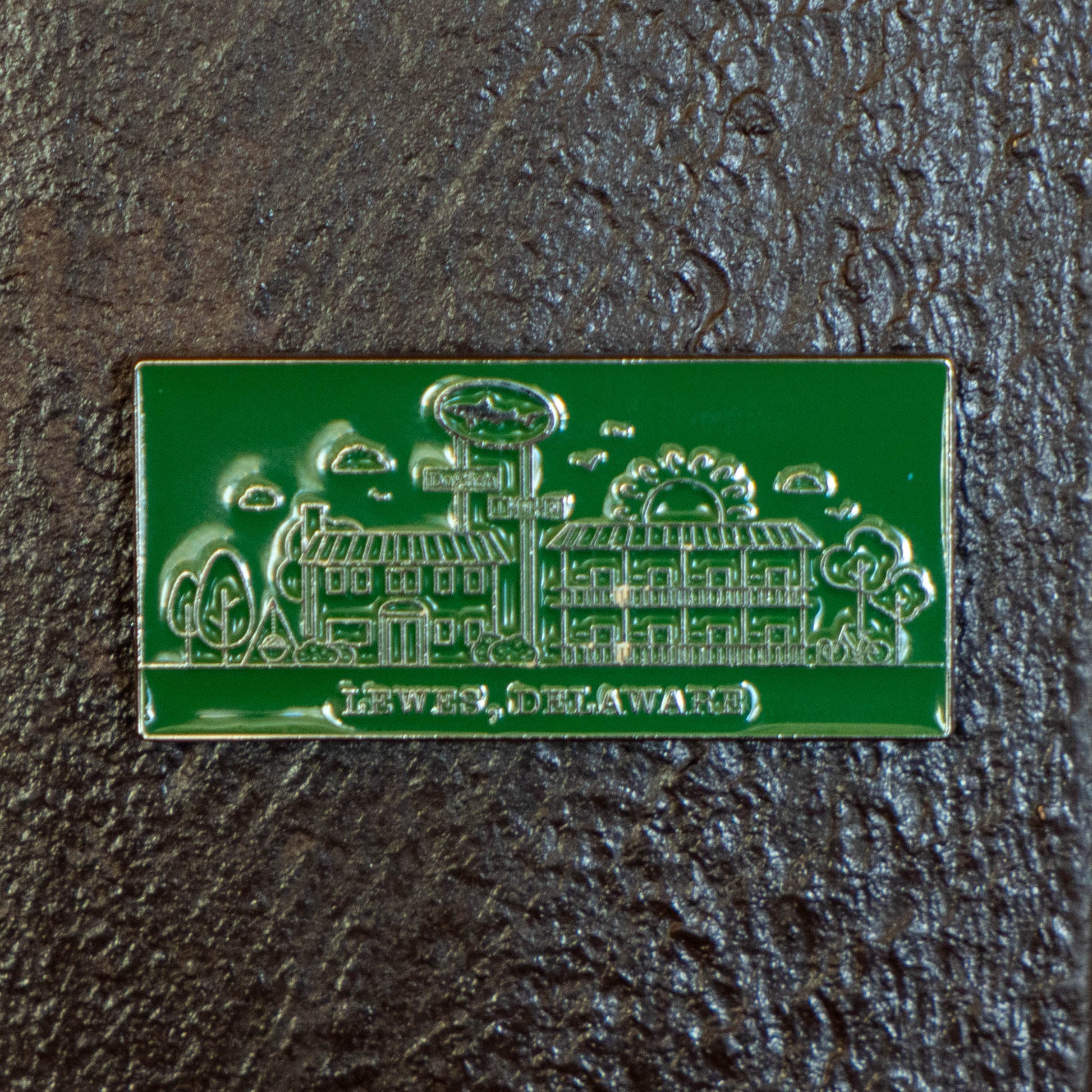 The Enamel Inn Magnet by Arrasmith Promotions LLC is a green enamel magnet featuring buildings, trees, clouds, and sun with "LEWES DELAWARE" text—perfect for Lewes or Dogfish Head fans—shown on a textured dark surface.