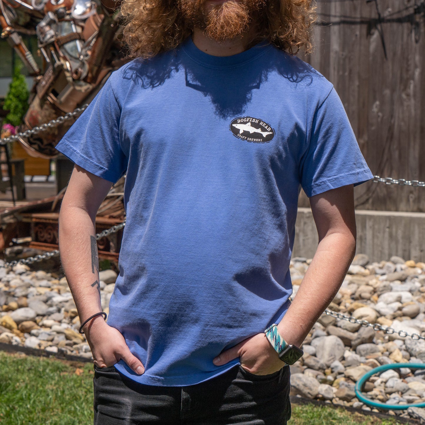 A person with curly hair and a beard stands outdoors wearing a FLO Blue Treehouse Short Sleeve Tee Shirt by GO WEST T-SHIRT COMPANY. Rocks, a chain fence, and greenery complete the background.