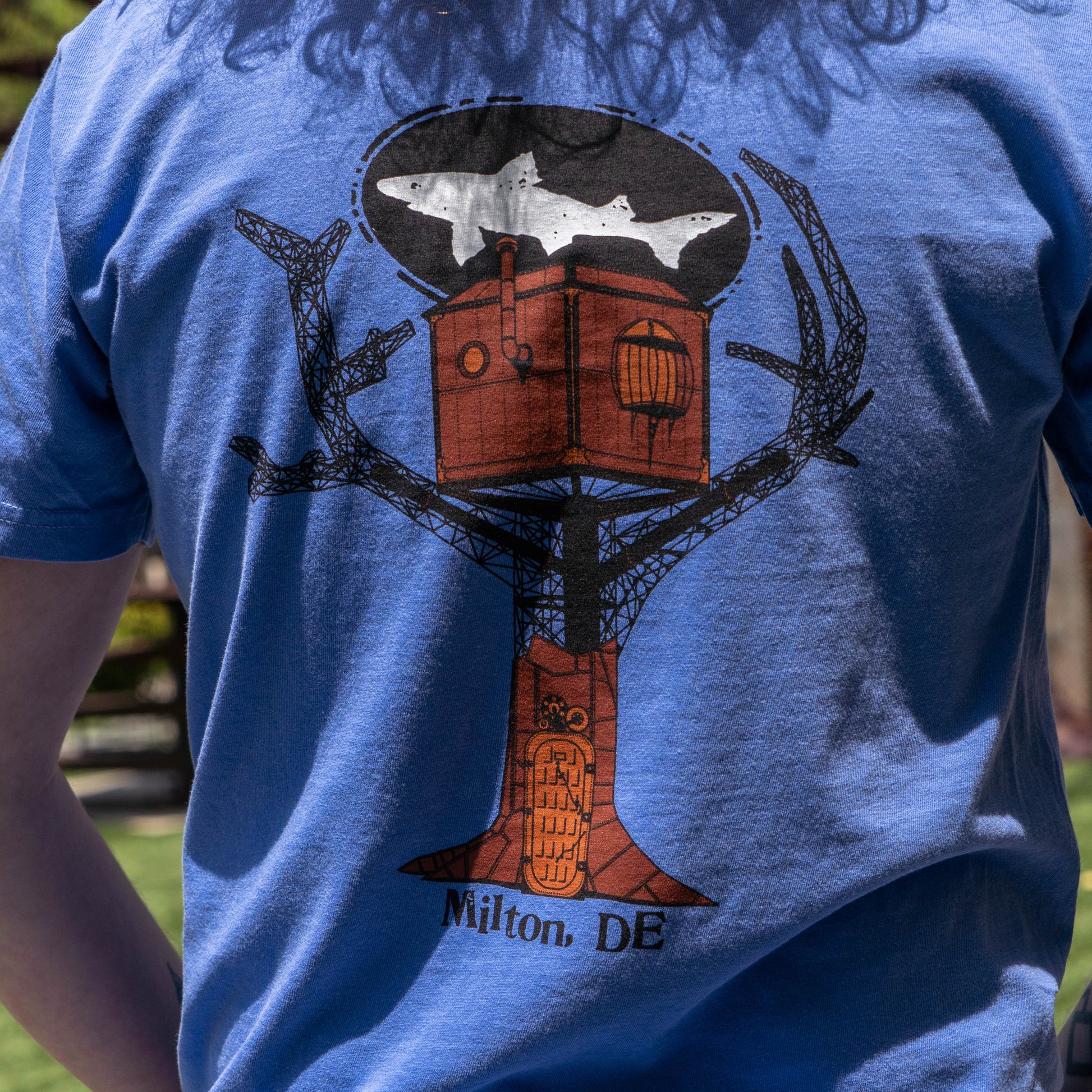 Someone wearing the FLO Blue Treehouse Short Sleeve Tee Shirt by GO WEST T-SHIRT COMPANY, which features an illustrated treehouse with a shark in an oval above and “Milton, DE” text at the bottom, outdoors on a sunny day.