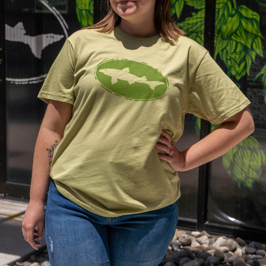A person stands outdoors in a sage green Fern Core Logo Tee Shirt by GO WEST T-SHIRT COMPANY, featuring an oval shark graphic, paired with blue jeans. Their left hand rests on their hip, and their face is partially out of frame.