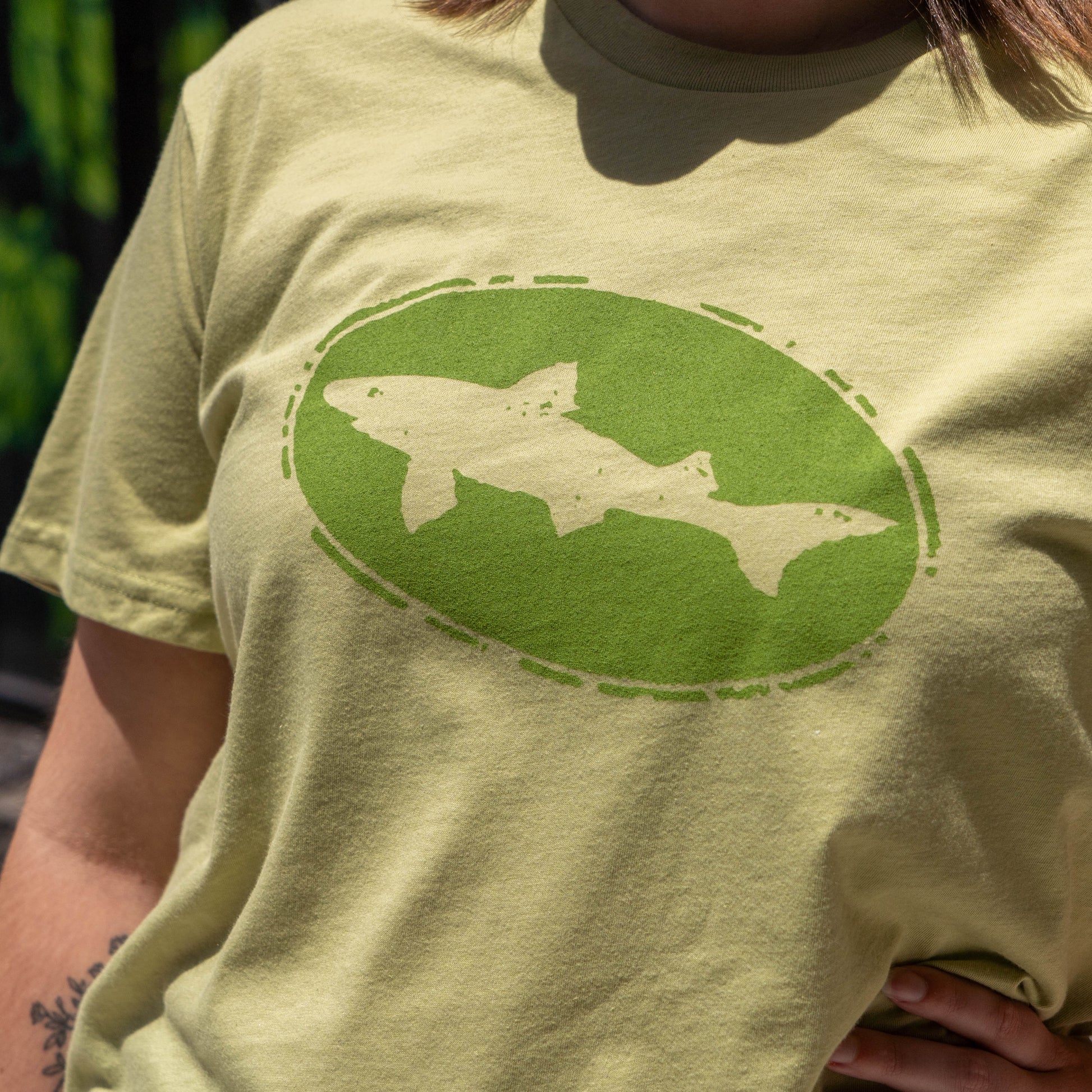 Someone wears a Fern Core Logo Tee Shirt by GO WEST T-SHIRT COMPANY in sage green, featuring a large stylized fish silhouette inside an oval on the chest. The unisex tee pops against the softly blurred background.