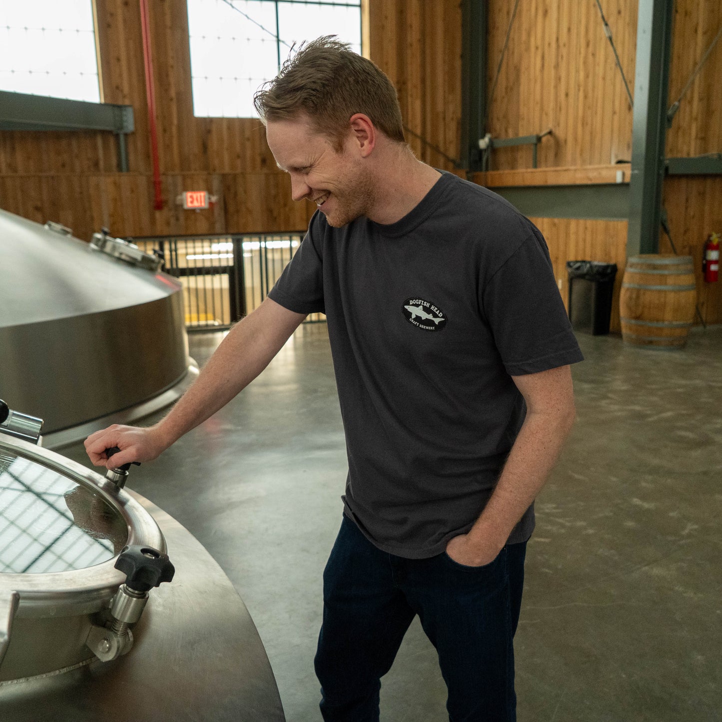 Man wearing a graphite gray Dogfish Head Delaware T‑shirt stands inside a brewery, shown from the front.