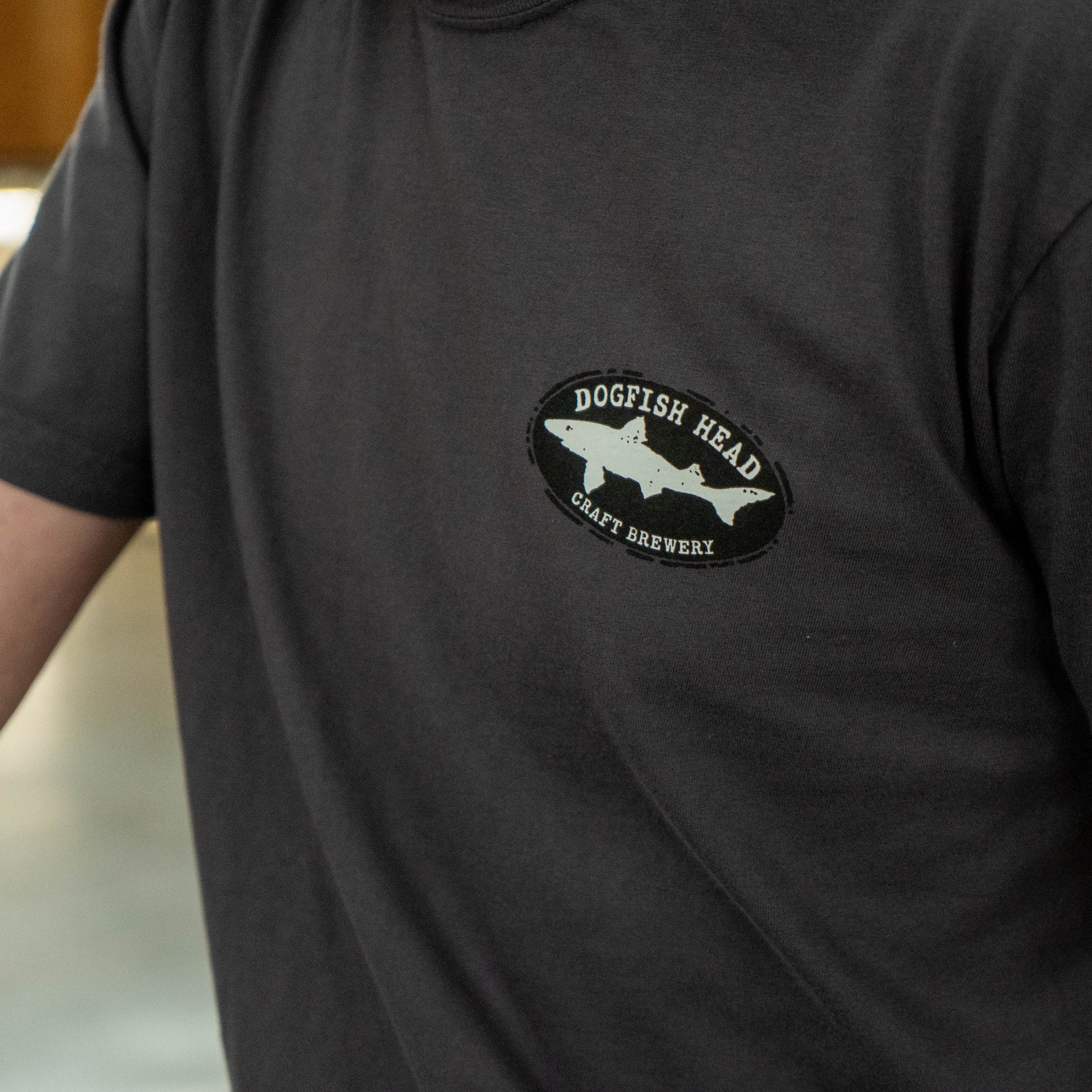 Close-up of the front chest logo on the graphite Dogfish Head T‑shirt, featuring the Dogfish Head Craft Brewery shark emblem