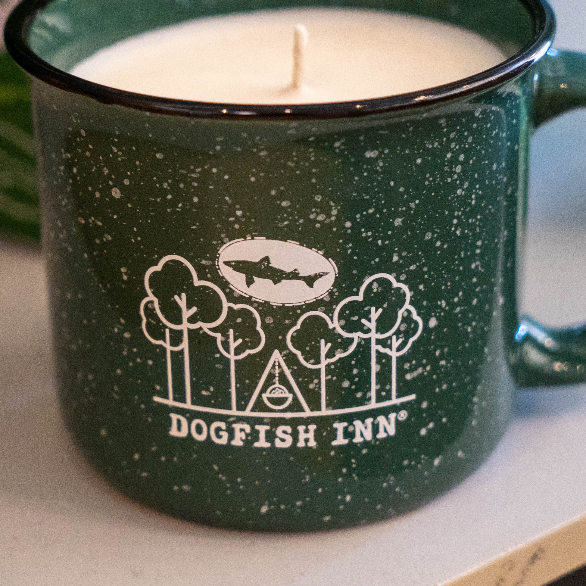The INN Camp Cup Candle by Dogfish Head features a forest green mug with white illustrations of trees, a tent, and the Dogfish Inn logo, filled with a white candle.