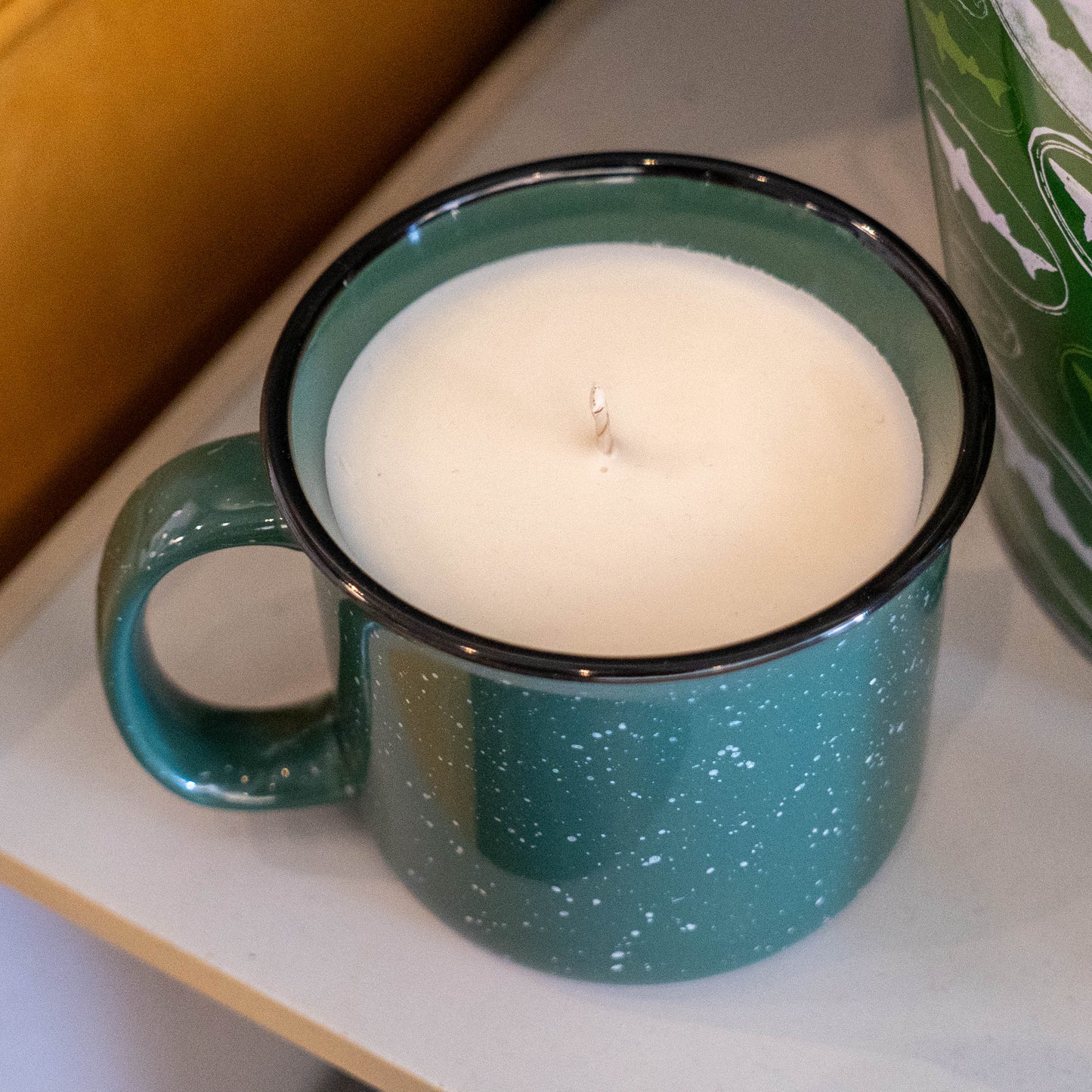 The INN Camp Cup Candle by Dogfish Head Brand features white wax with a single wick in a forest green ceramic mug with a handle and black rim, displayed on a white surface.