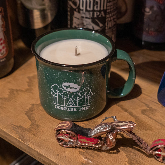 A Dogfish Head Brand INN Camp Cup Candle in forest green rests on a wooden surface next to a small metal motorcycle, with bottles in the background.