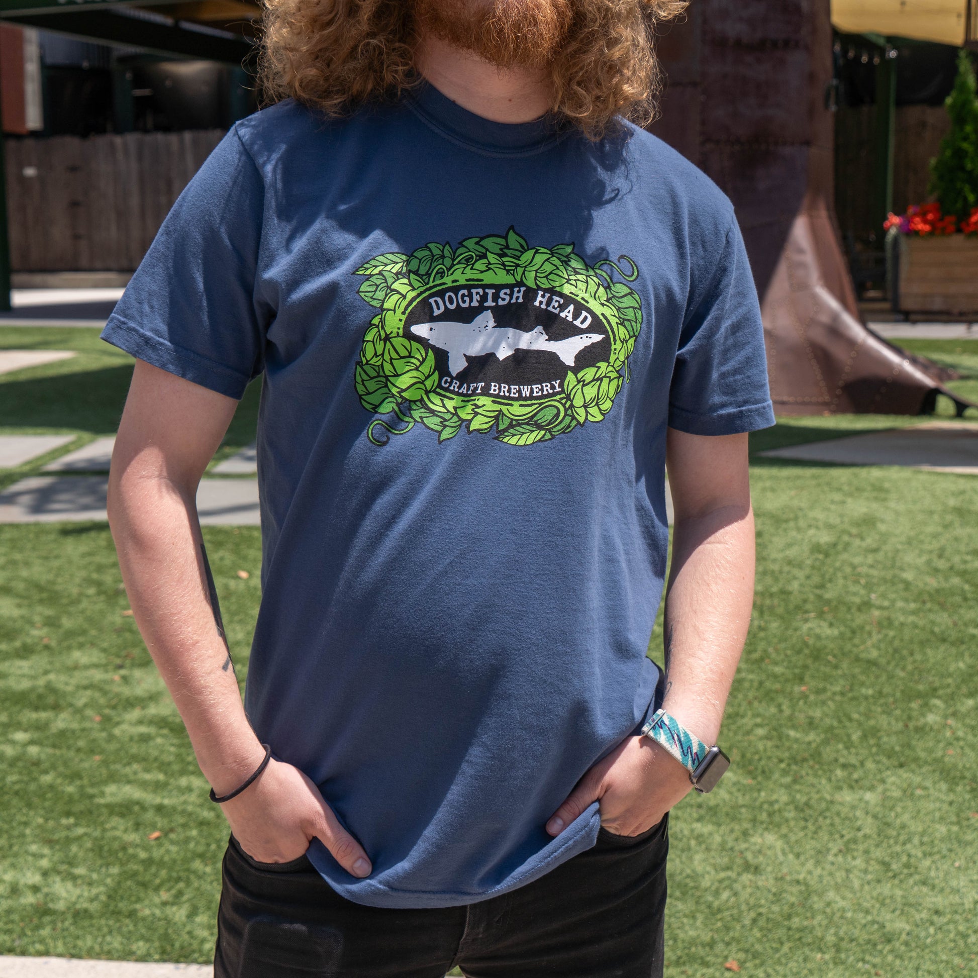 A curly-haired, bearded person stands outdoors wearing the GO WEST T-SHIRT COMPANY Midnight Got a Minute? Tee Shirt, holding its sides. Green grass and buildings in the background highlight this unique craft beer-inspired apparel.