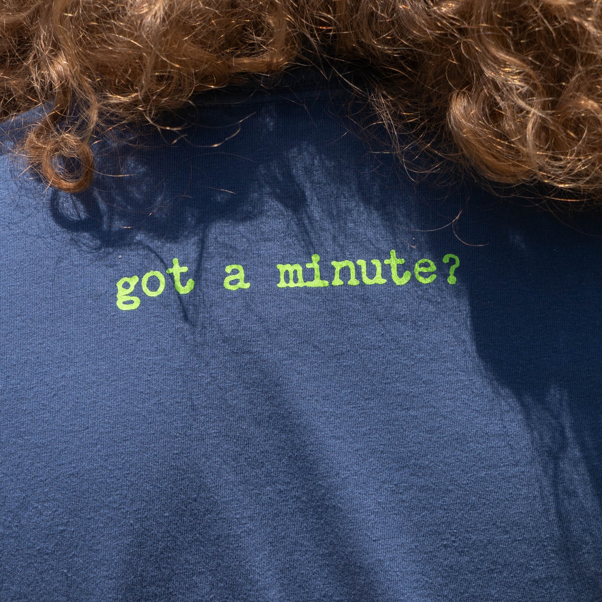 The Midnight Got a Minute? Tee Shirt by GO WEST T-SHIRT COMPANY features “got a minute?” in light green on the back—a dark blue craft beer tee ideal for fans and casual outings. Brown curly hair partially covers its upper back.