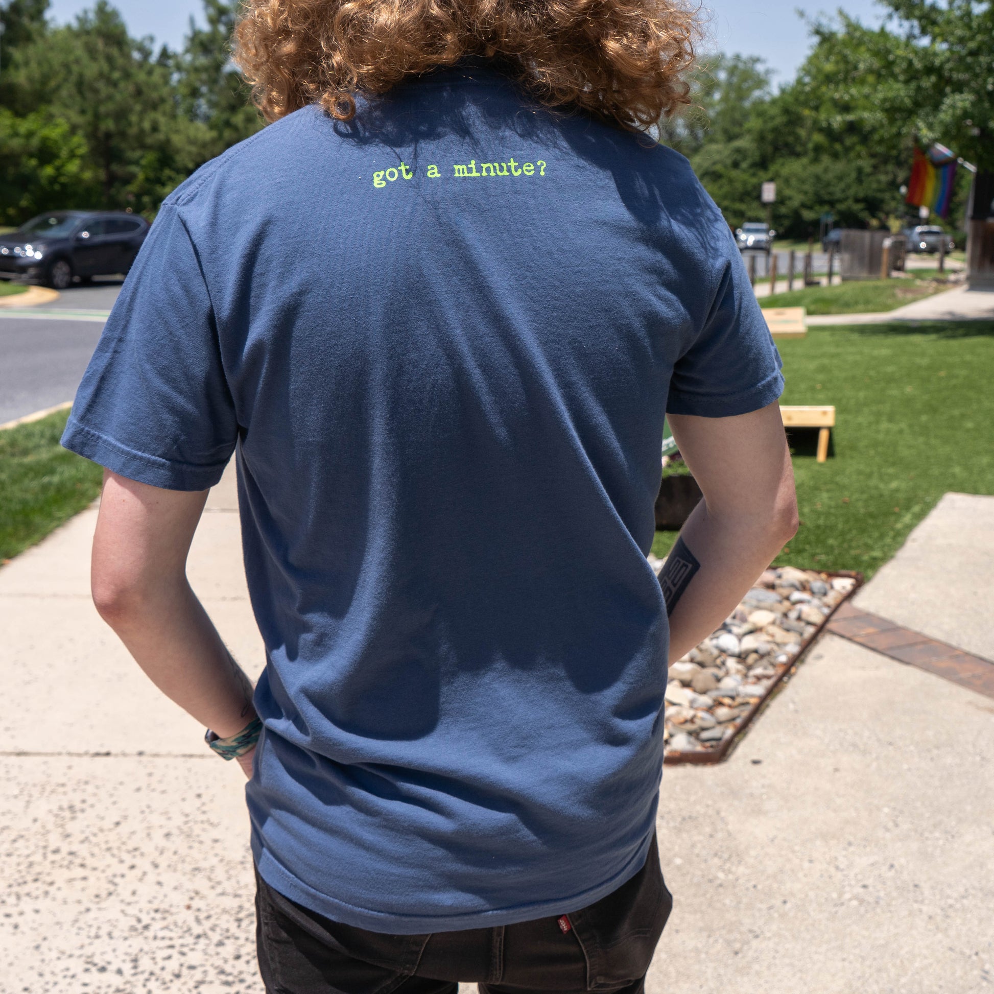 A person with curly hair stands outdoors in a GO WEST T-SHIRT COMPANY Midnight Got a Minute? Tee Shirt, featuring a blue hop graphic and "got a minute?" printed in green on the upper back, with trees and grass in the background.