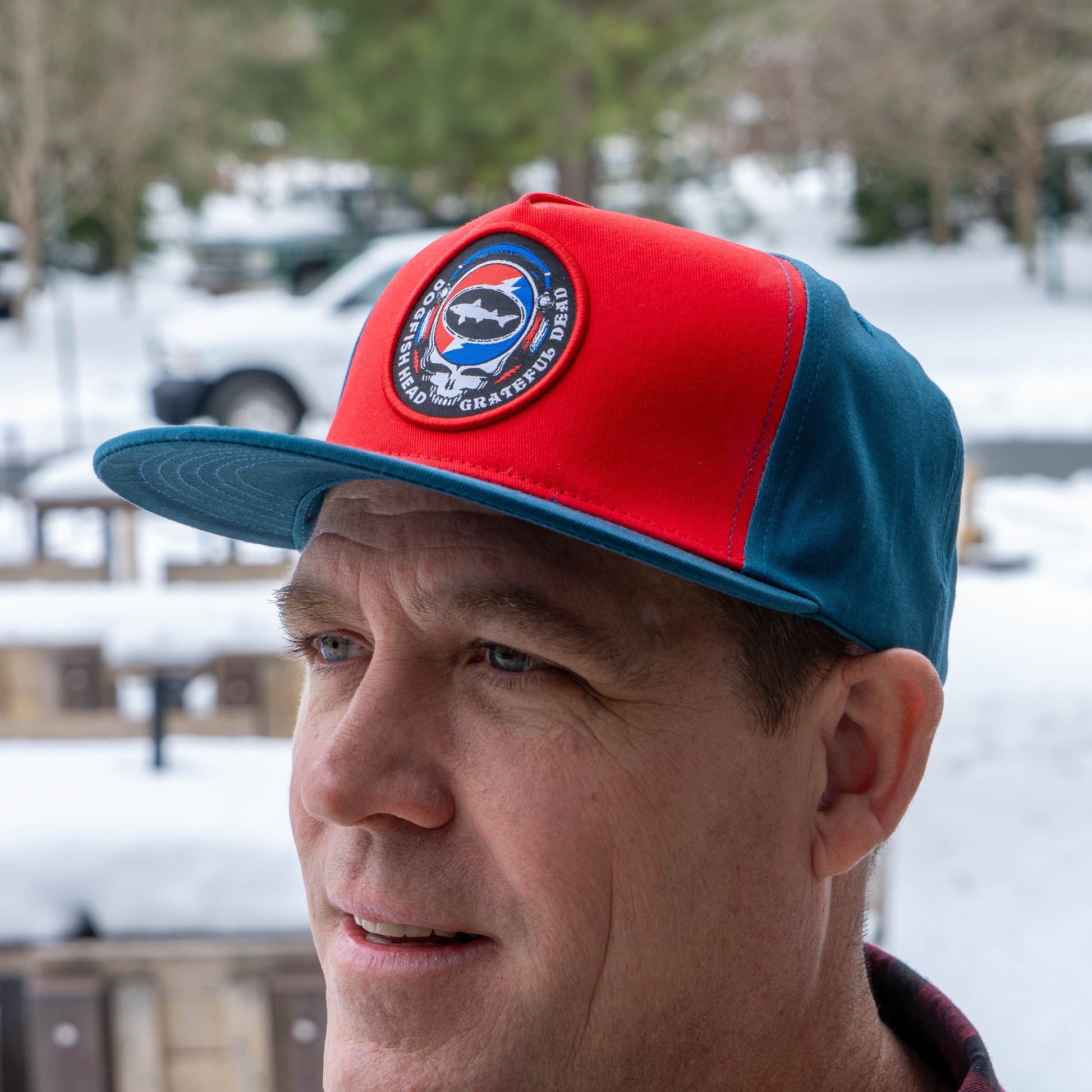 Man wearing Music Month Trucker Hat with a Dogfish Head x Grateful Dead logo. Designed with red front panels and cerulean blue back panels and brim. 
