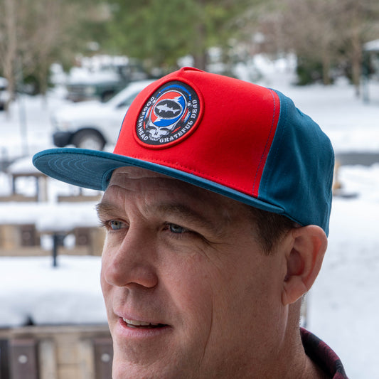 Man wearing Music Month Trucker Hat with a Dogfish Head x Grateful Dead logo. Designed with red front panels and cerulean blue back panels and brim. 