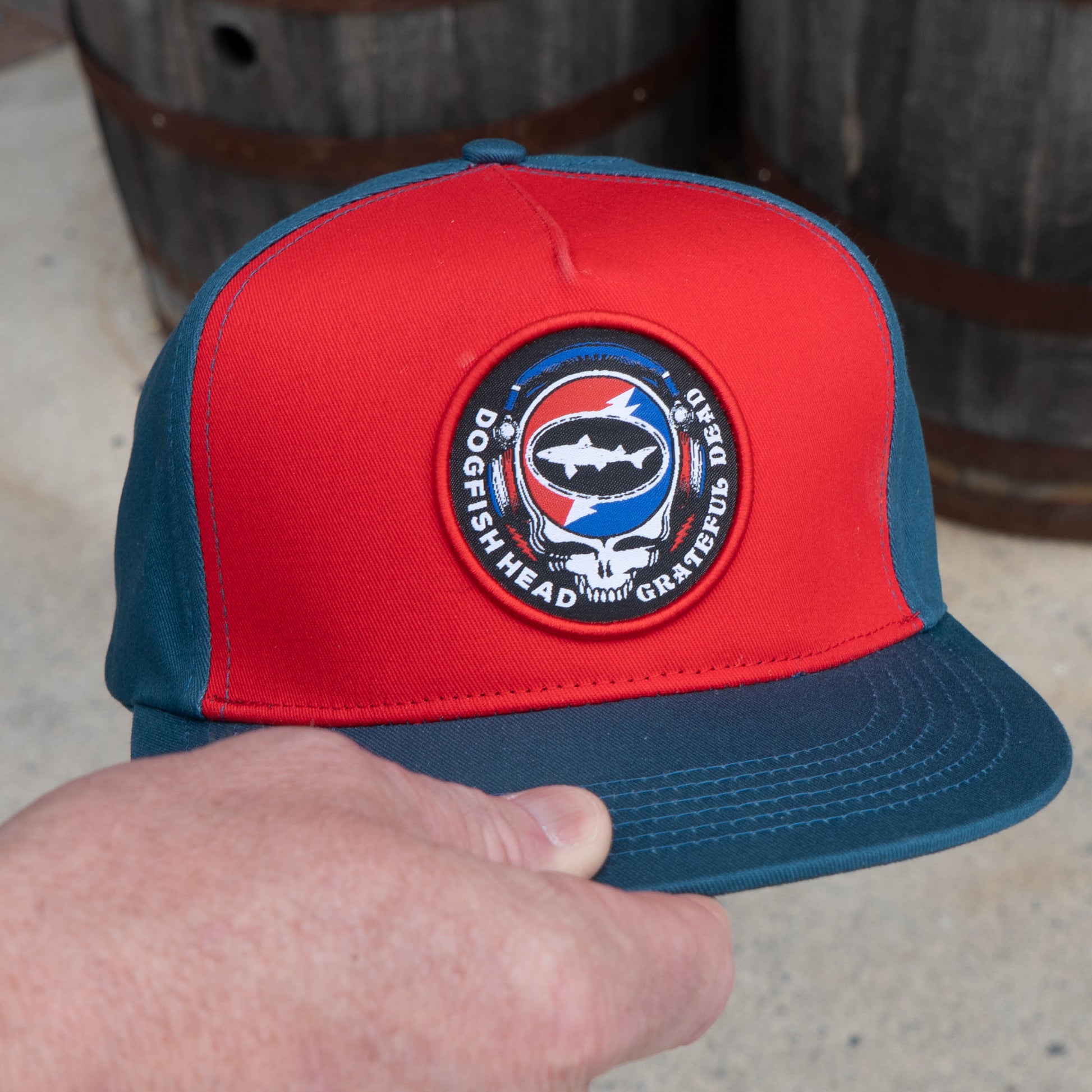 Hand holding the Music Month Trucker Hat with a Dogfish Head x Grateful Dead logo. Designed with red front panels and cerulean blue back panels and brim. 