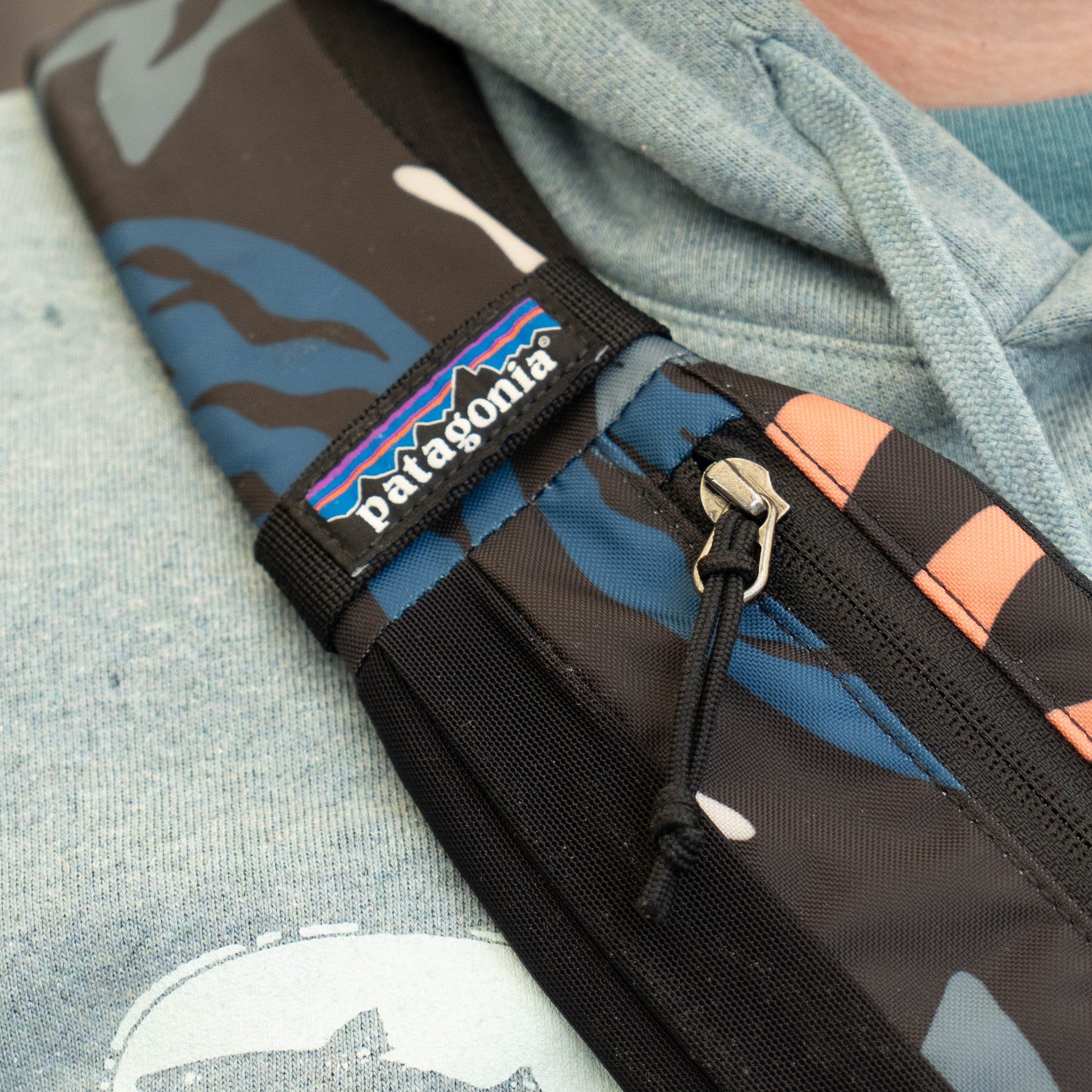 Close-up of Patagonia Atom Sling 8L strap with logo tag and zipper detail, durable camo fabric crossbody daypack design