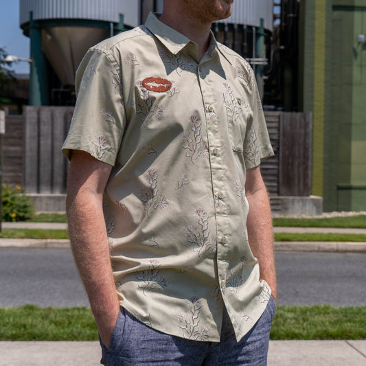 A man stands outdoors with his hands in his pockets, wearing a Patagonia M Brown Go To Shirt by Patagonia—a light, organic cotton short-sleeve button-up. Industrial tanks and buildings are visible in the background.