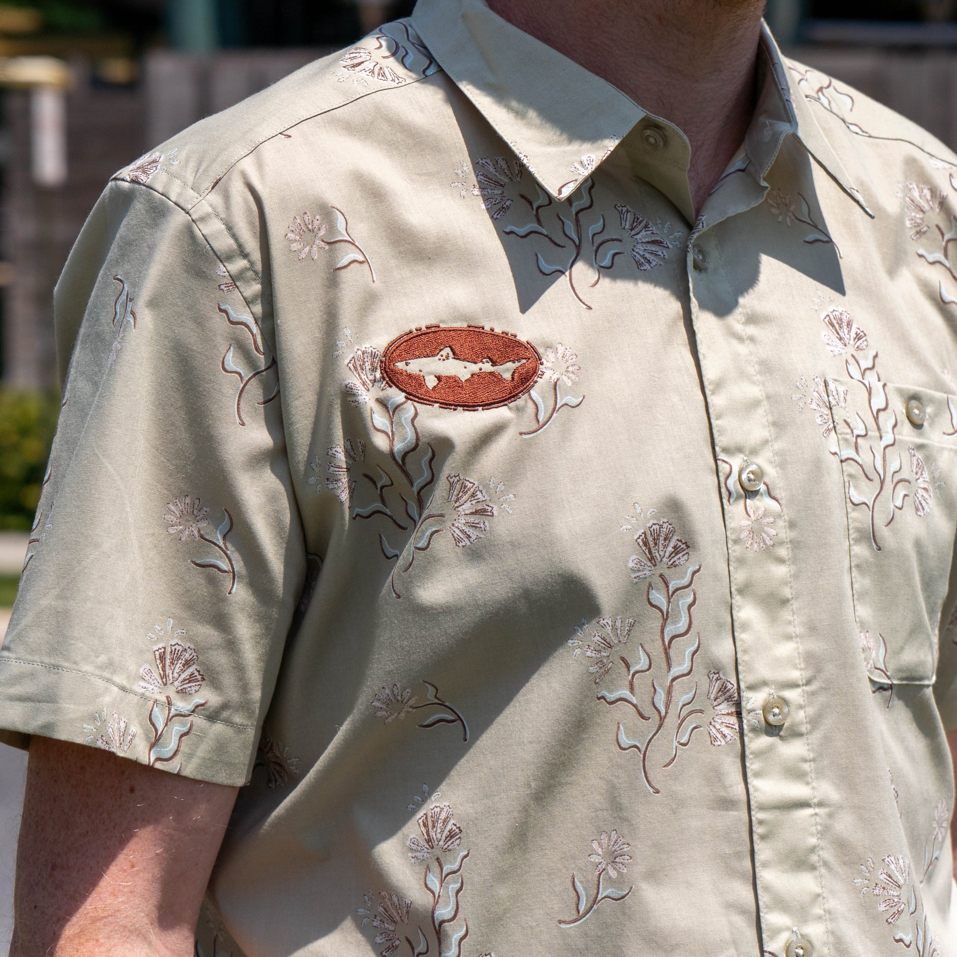 Someone is wearing a Patagonia M Brown Go To Shirt—an organic cotton, light beige short-sleeve button-up with a floral pattern and a brown oval patch featuring a white fish on the chest. The photo is taken outdoors.