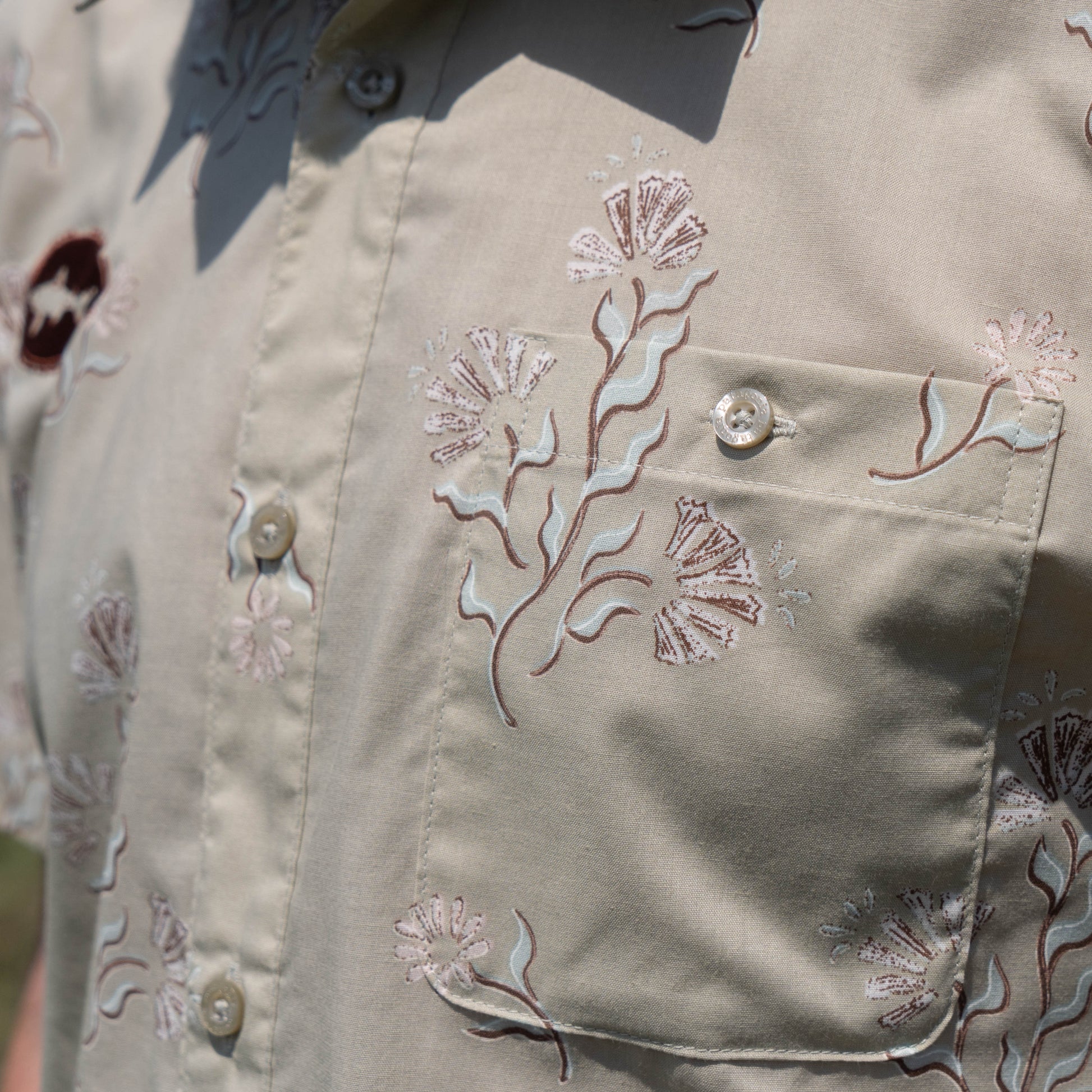 Close-up of the Patagonia M Brown Go To Shirt, a beige short-sleeve organic cotton button-up by Patagonia, featuring floral embroidery in light pink and brown, a front pocket, and several buttons.