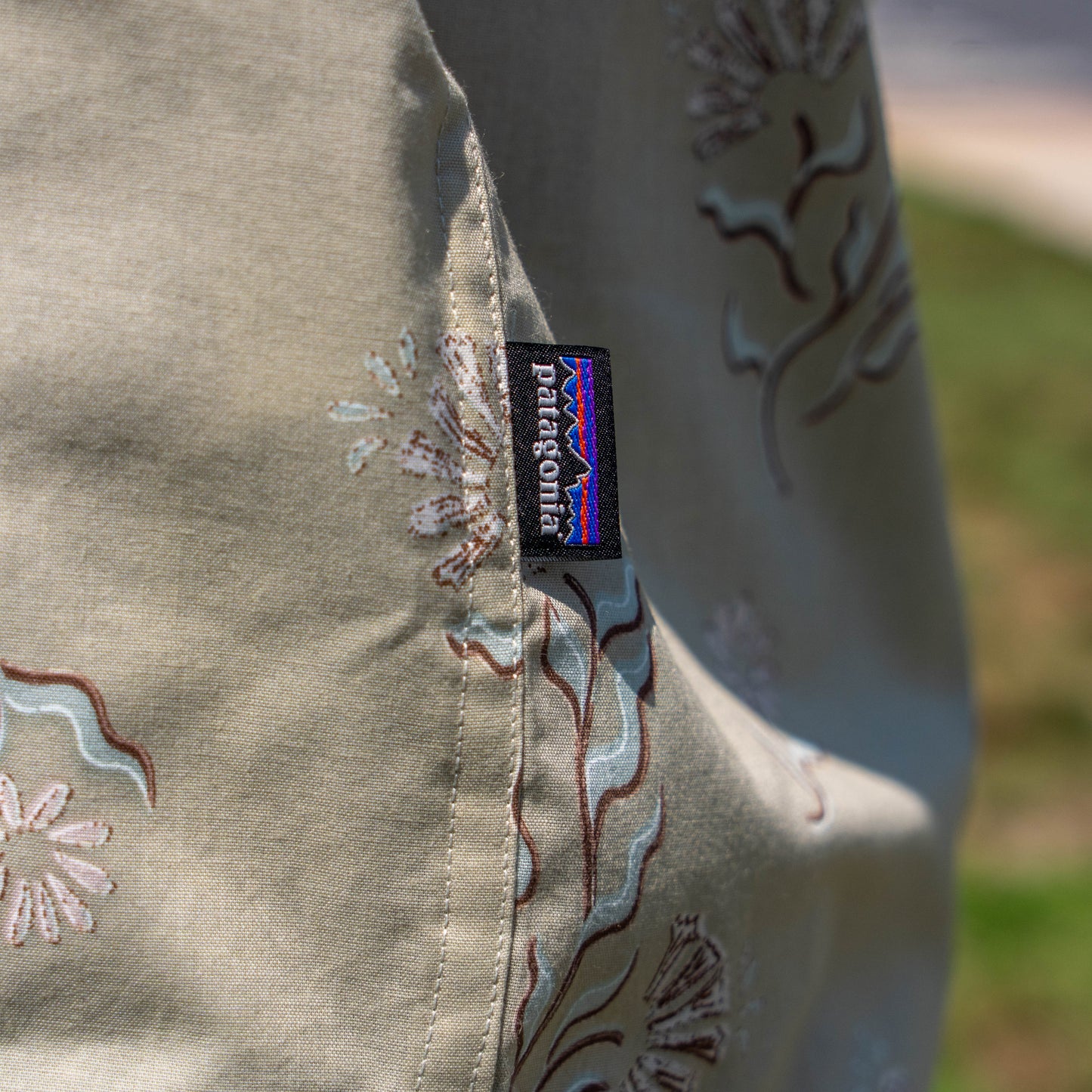 Close-up of the Patagonia M Brown Go To Shirt with a floral pattern and a small black Patagonia tag on the seam; background shows blurred grass and sidewalk.