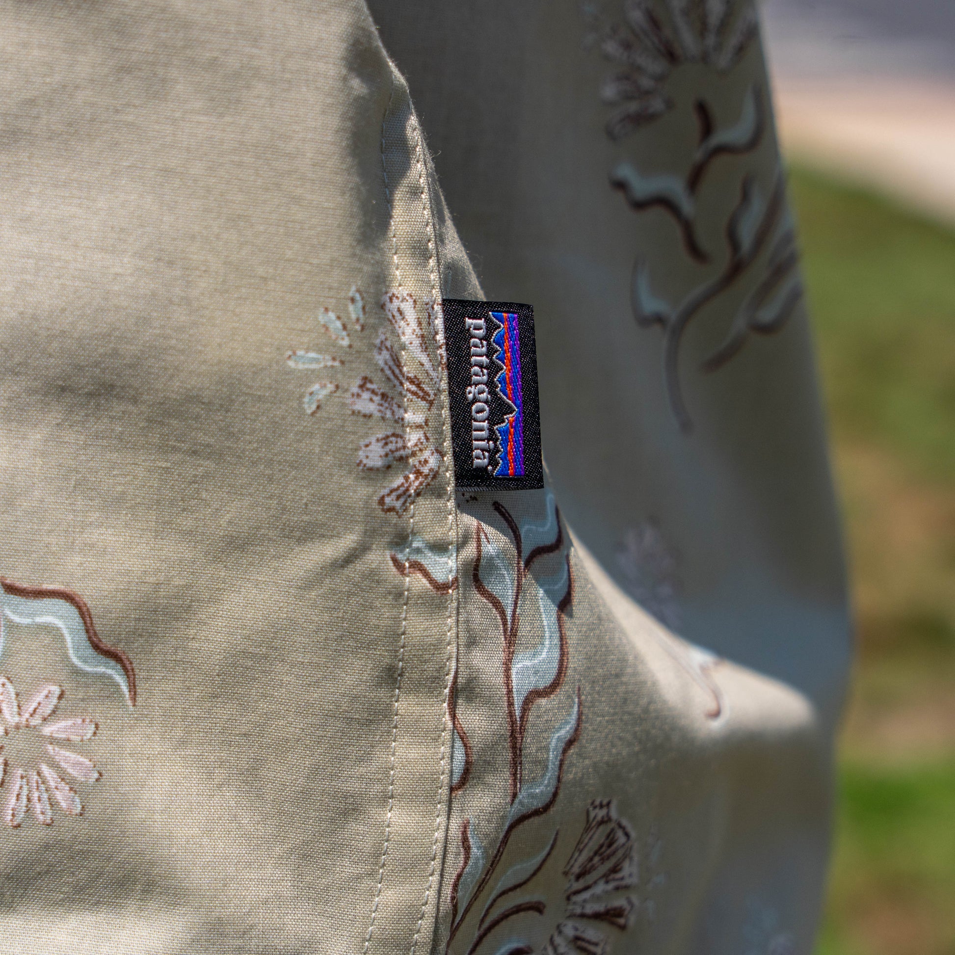 Close-up of the Patagonia M Brown Go To Shirt with a floral pattern and a small black Patagonia tag on the seam; background shows blurred grass and sidewalk.