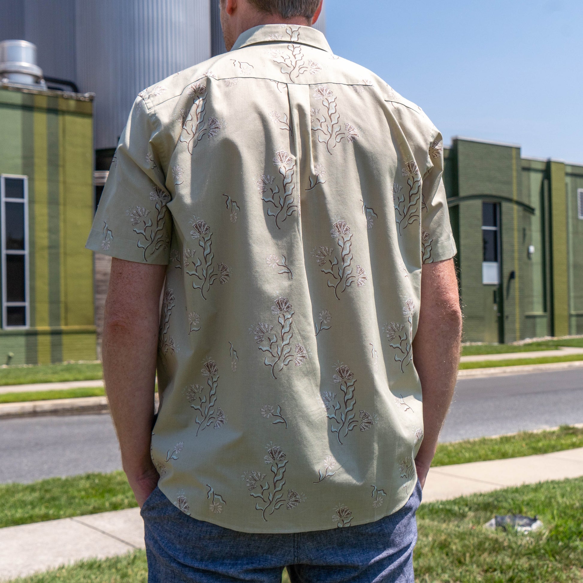 Someone stands outdoors with their back to the camera, wearing a Patagonia M Brown Go To Shirt. A green industrial-style building and sidewalk are visible in the background.