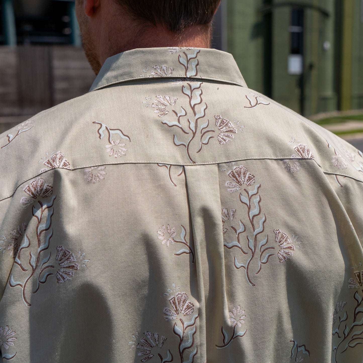 A person wearing the Patagonia M Brown Go To Shirt by Patagonia stands outdoors facing away, with buildings and grass blurred in the background.
