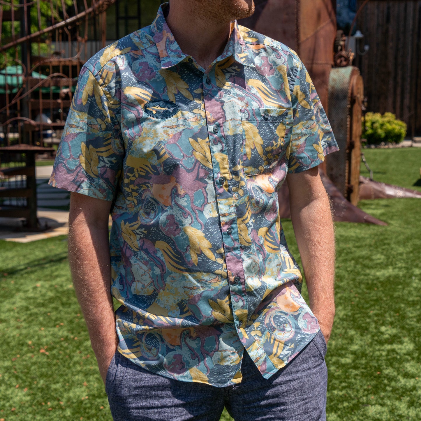 A man stands outdoors on green grass with his hands in his pockets, wearing blue-gray pants and the Patagonia M Floral Pattern Go To Shirt by Patagonia.