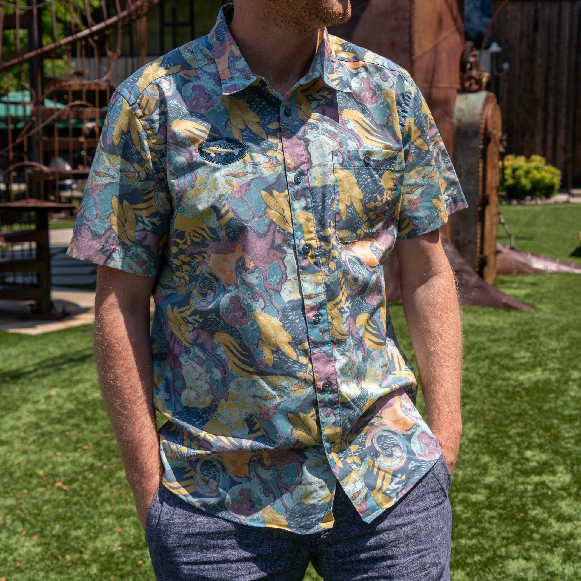A man stands outdoors on green grass with his hands in his pockets, wearing blue-gray pants and the Patagonia M Floral Pattern Go To Shirt by Patagonia.