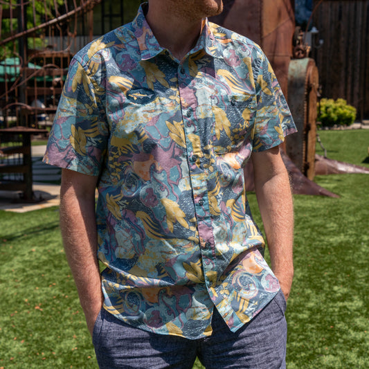 A man stands outdoors on green grass with his hands in his pockets, wearing blue-gray pants and the Patagonia M Floral Pattern Go To Shirt by Patagonia.