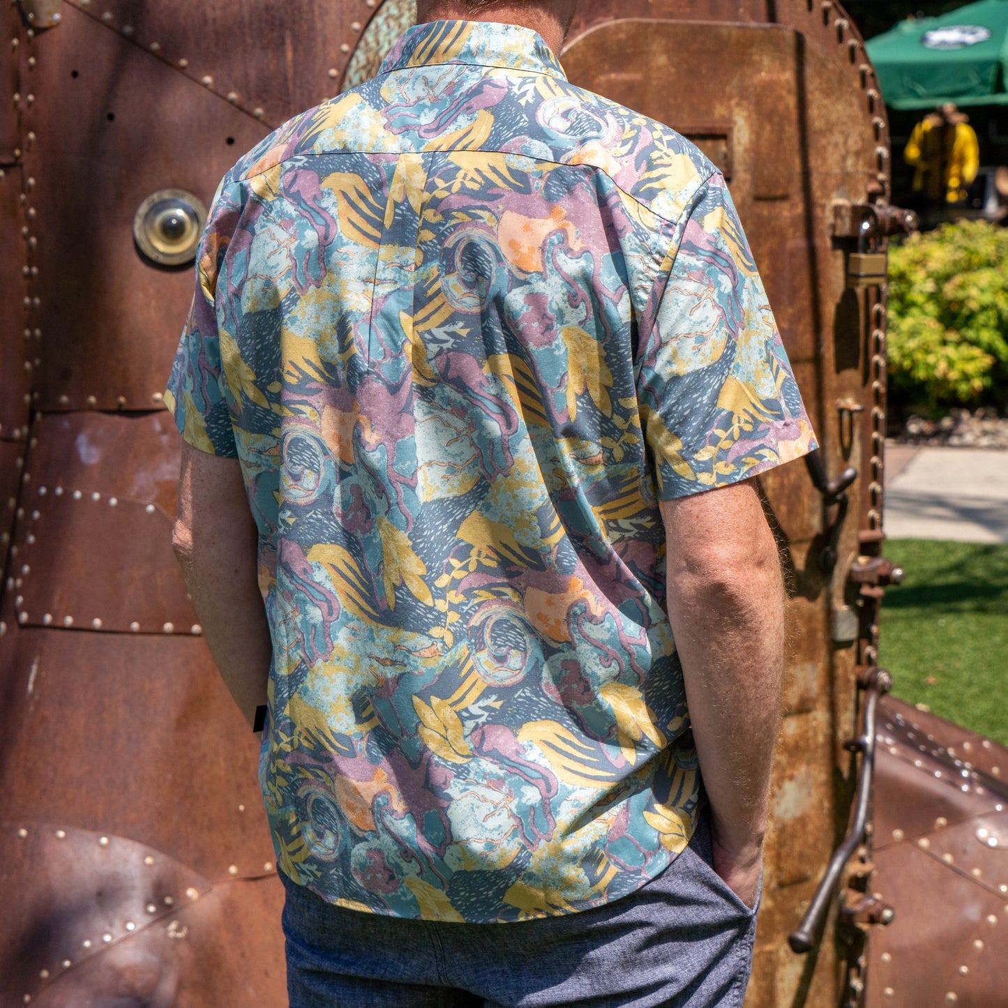 A person stands outdoors with their back to the camera, wearing a Patagonia M Floral Pattern Go To Shirt by Patagonia, featuring vibrant floral designs, beside a large metallic steampunk-style structure.