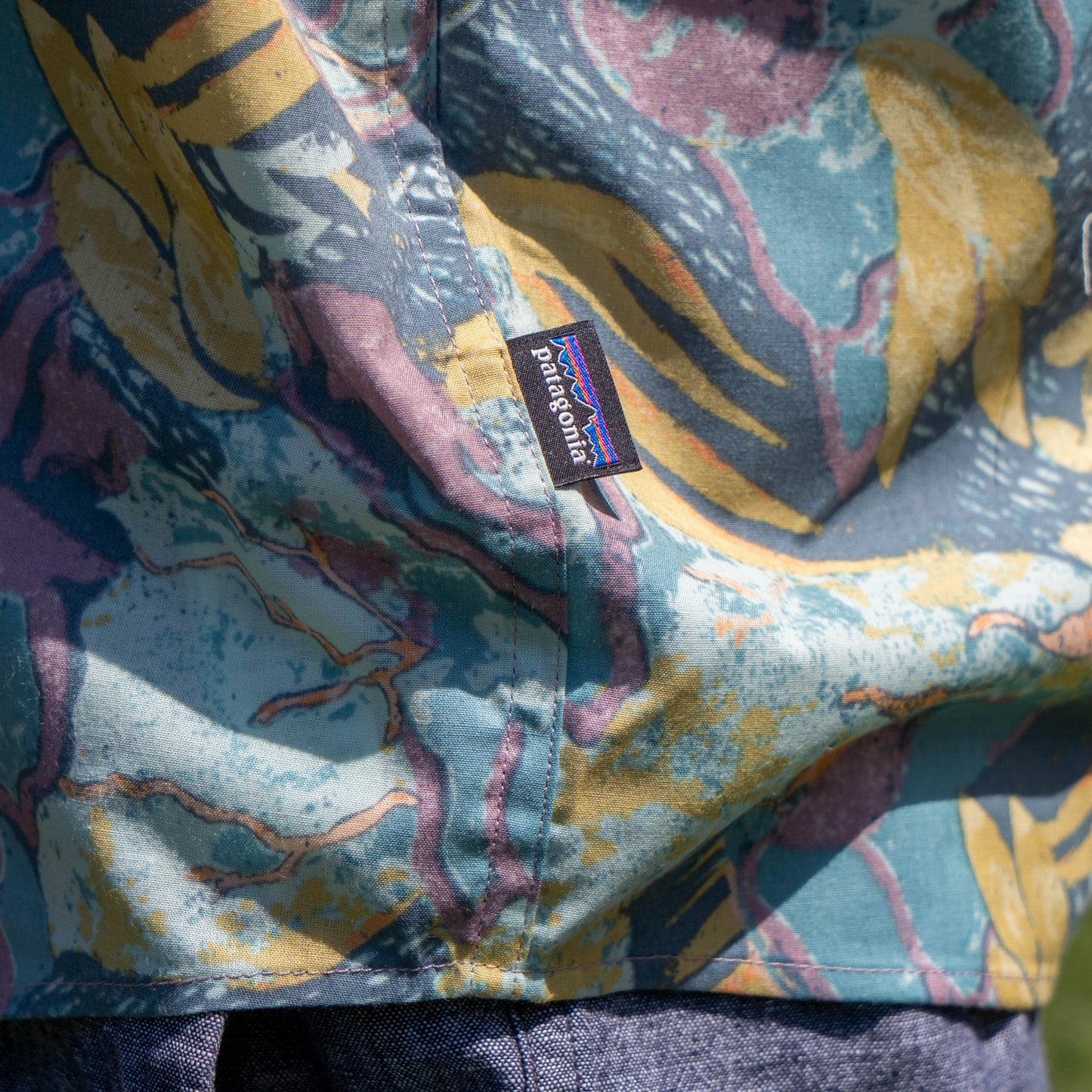 Close-up of the Patagonia M Floral Pattern Go To Shirt—a lightweight, multicolored button-up with abstract patterns and a brand tag by the side seam; blue denim pants appear below.
