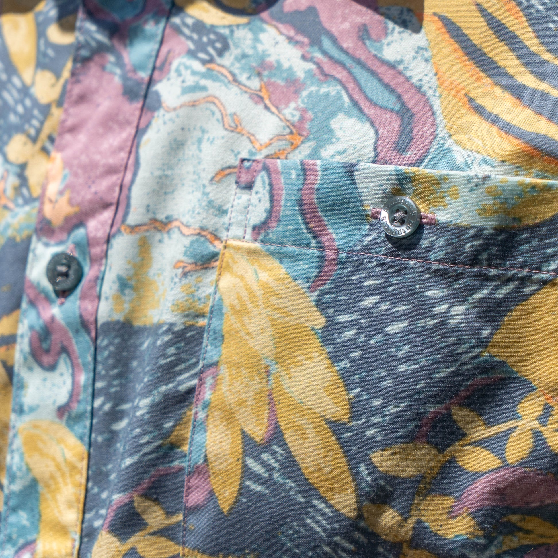 A close-up of the Patagonia M Floral Pattern Go To Shirt by Patagonia shows its abstract blue, yellow, pink, and white leaf and wavy designs on organic cotton. The lightweight shirt features a pocket, buttons, and sunlight highlighting the textured fabric.