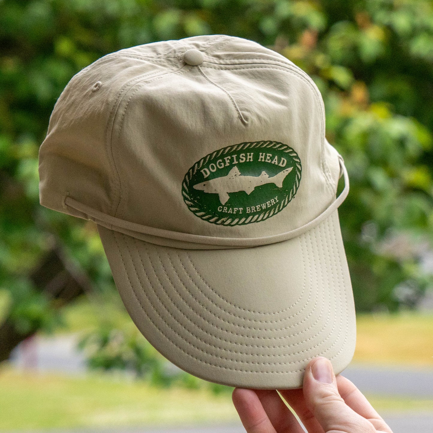 Hand holding patagonia Dogfish head spoonbill merganzer tan hat with a green Dogfish Head Craft Brewery logo in the center and a double duty adjustable chin strap 