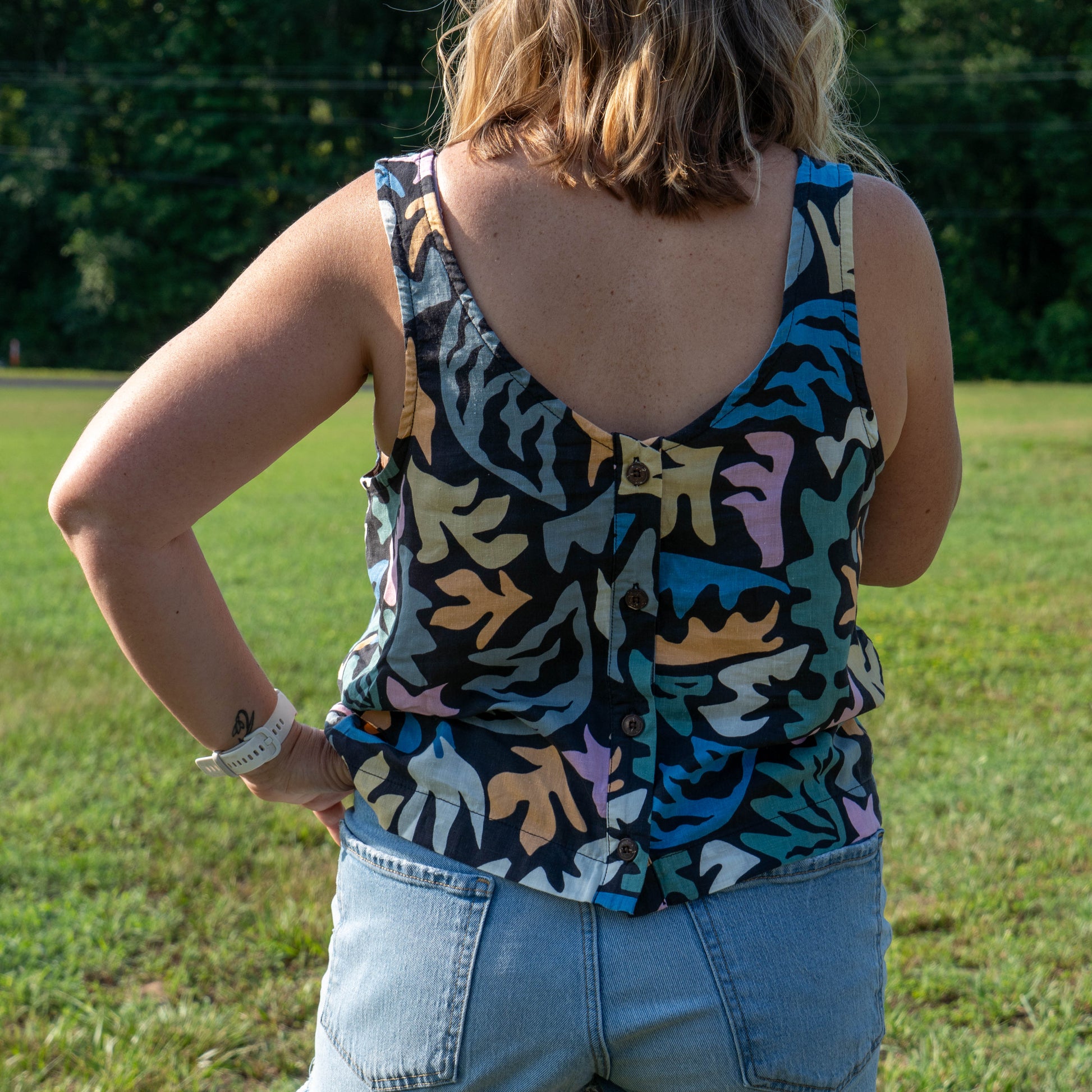 A person stands outdoors with their back to the camera, wearing a Patagonia W Garden Island Tank Top and light blue jean shorts. The background shows green grass and trees.